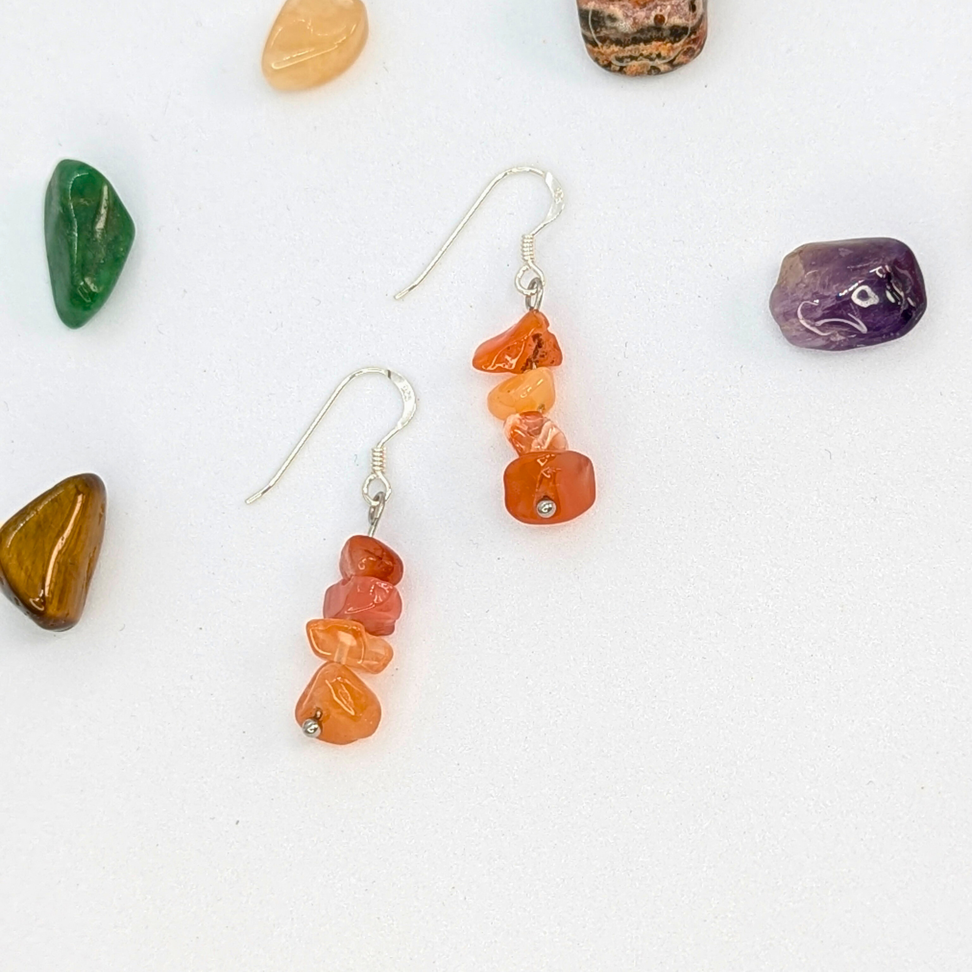 Silver Sunbird Carnelian Agate Earrings with orange gemstone chips and sterling silver dangle hooks are shown on a white surface, surrounded by polished green, purple, brown, and tan stones.