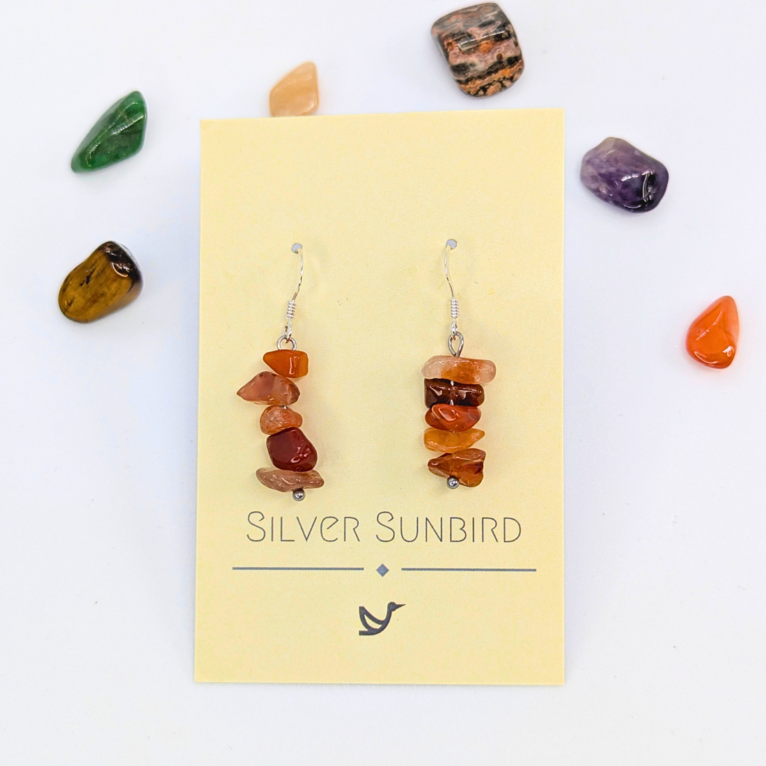 Silver Sunbird’s Carnelian Agate Earrings, featuring stacked reddish-brown stones, are displayed on a branded card and surrounded by various polished gemstones against a white background.