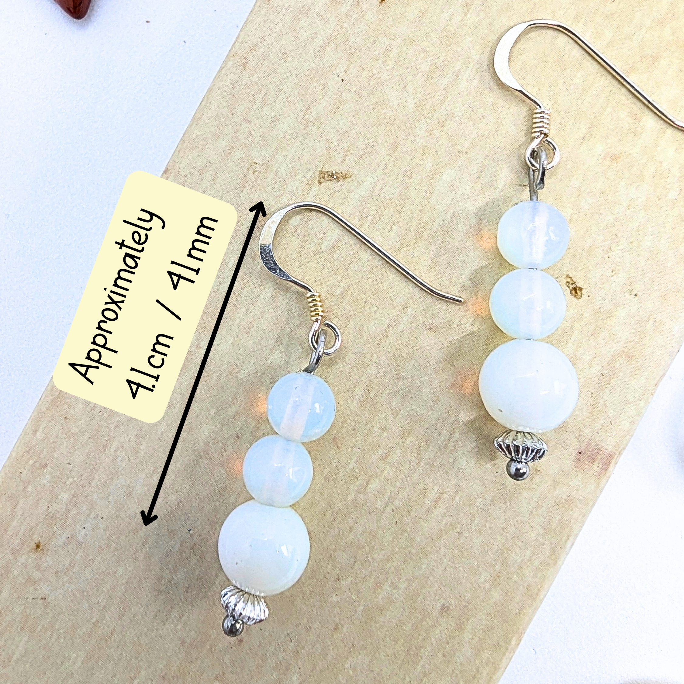 The Opal Dangle Earrings by Silver Sunbird have graduated white beads on silver hooks, measuring about 4.1 cm or 41 mm, and are displayed on a wooden surface.