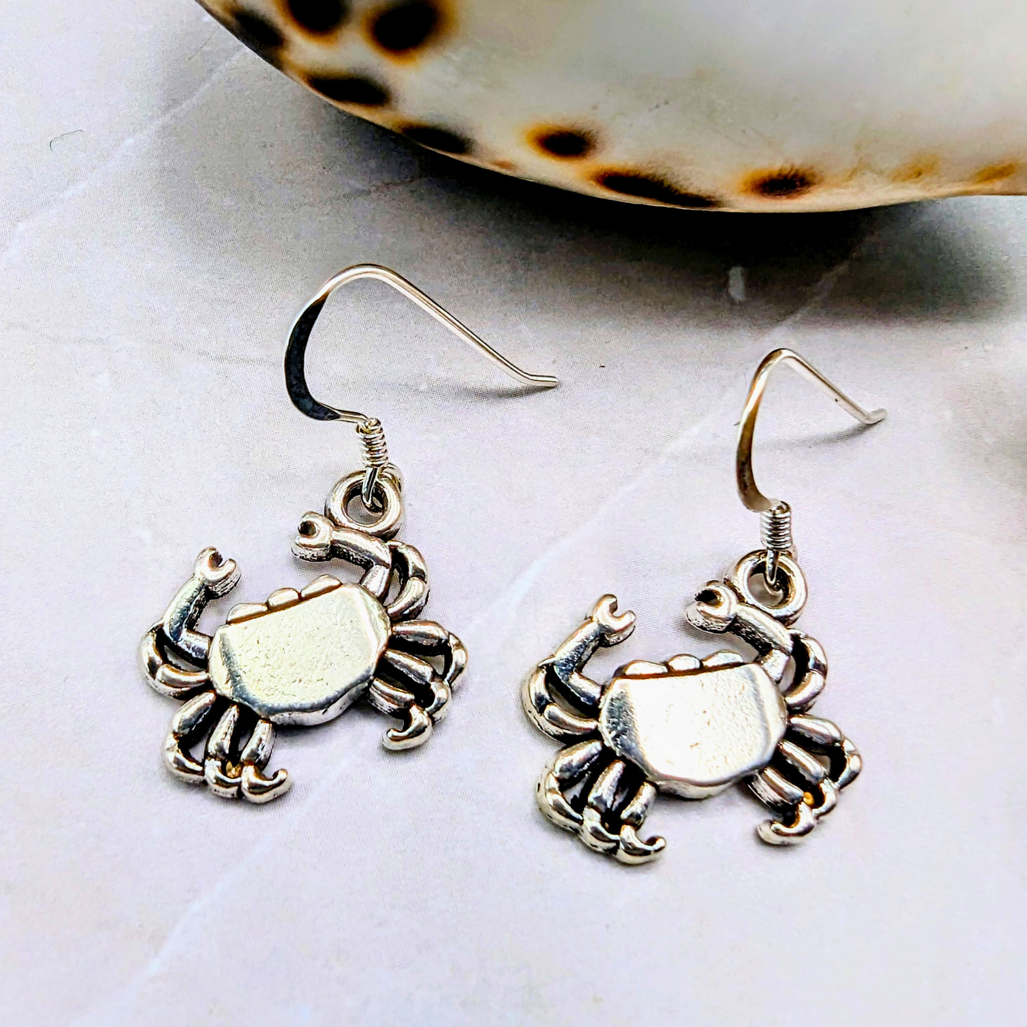 Coastal Crab Earrings by Silver Sunbird, crafted in sterling silver, are shown on a light marble surface with a spotted seashell partially visible in the background.
