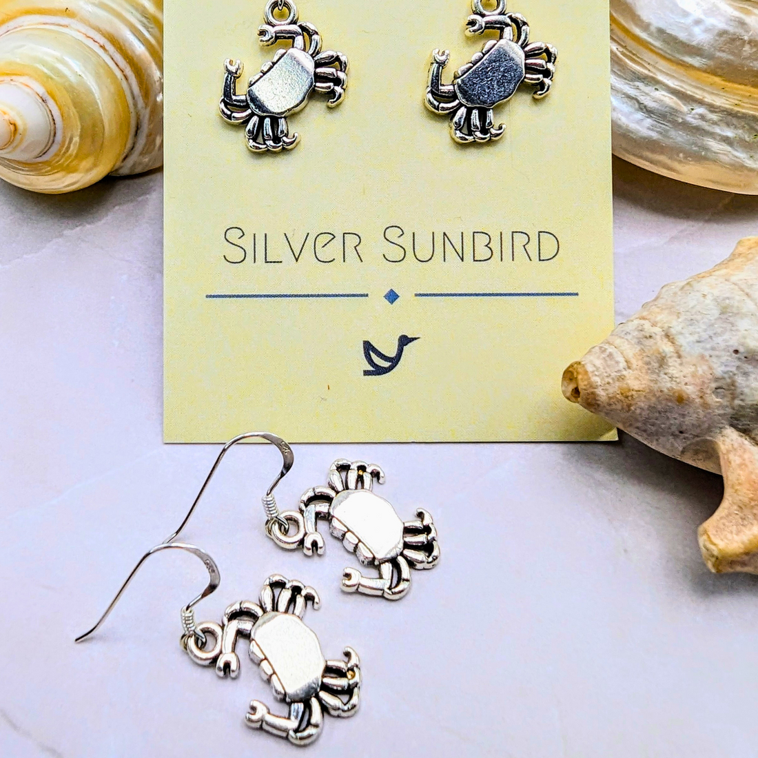 Coastal Crab Earrings by Silver Sunbird are shown on a branded card, surrounded by seashells on a light surface.