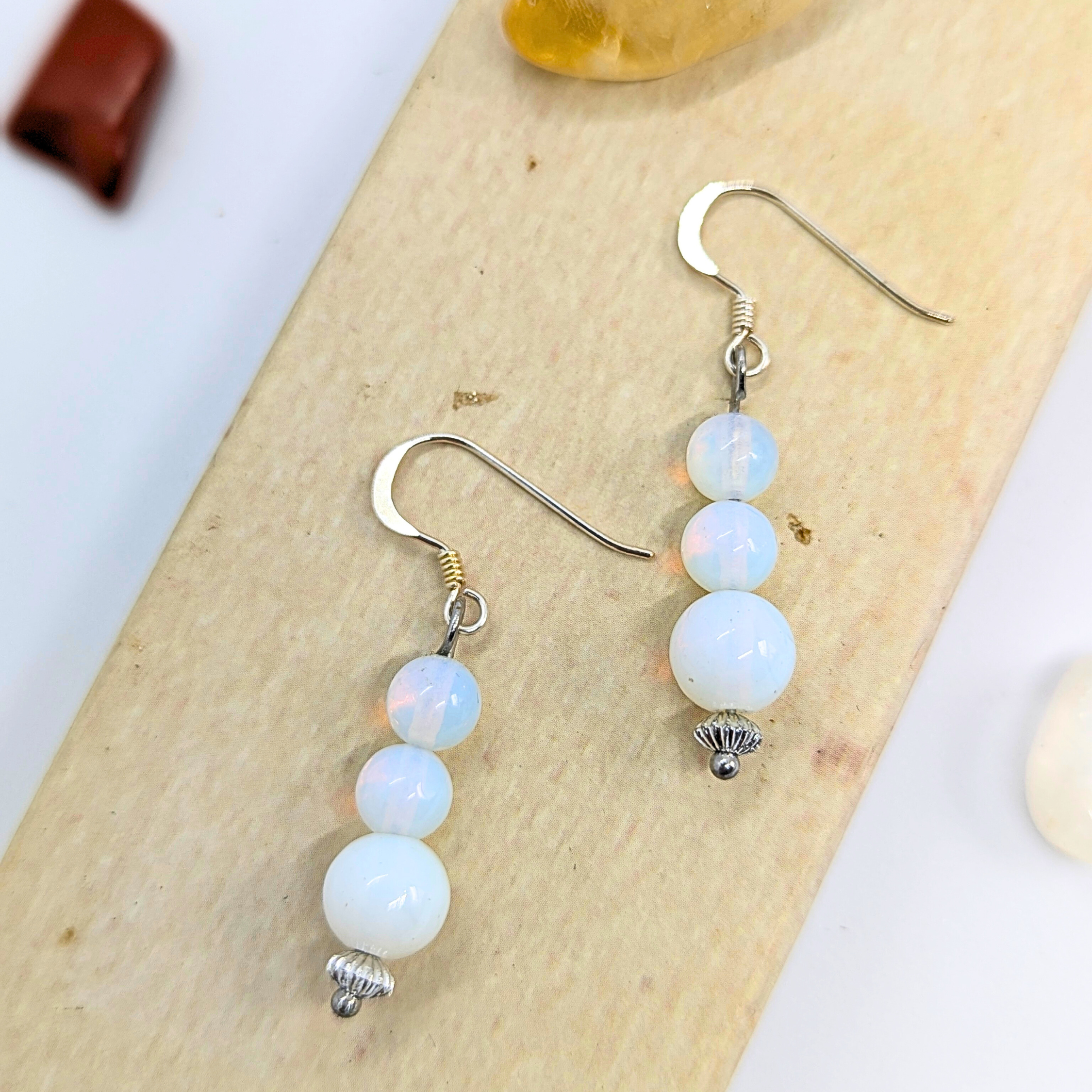 Opal Dangle Earrings by Silver Sunbird feature handmade designs with stacked opal-like iridescent beads in a subtle color range, silver hooks, and ornate bead caps on a light wood backdrop.