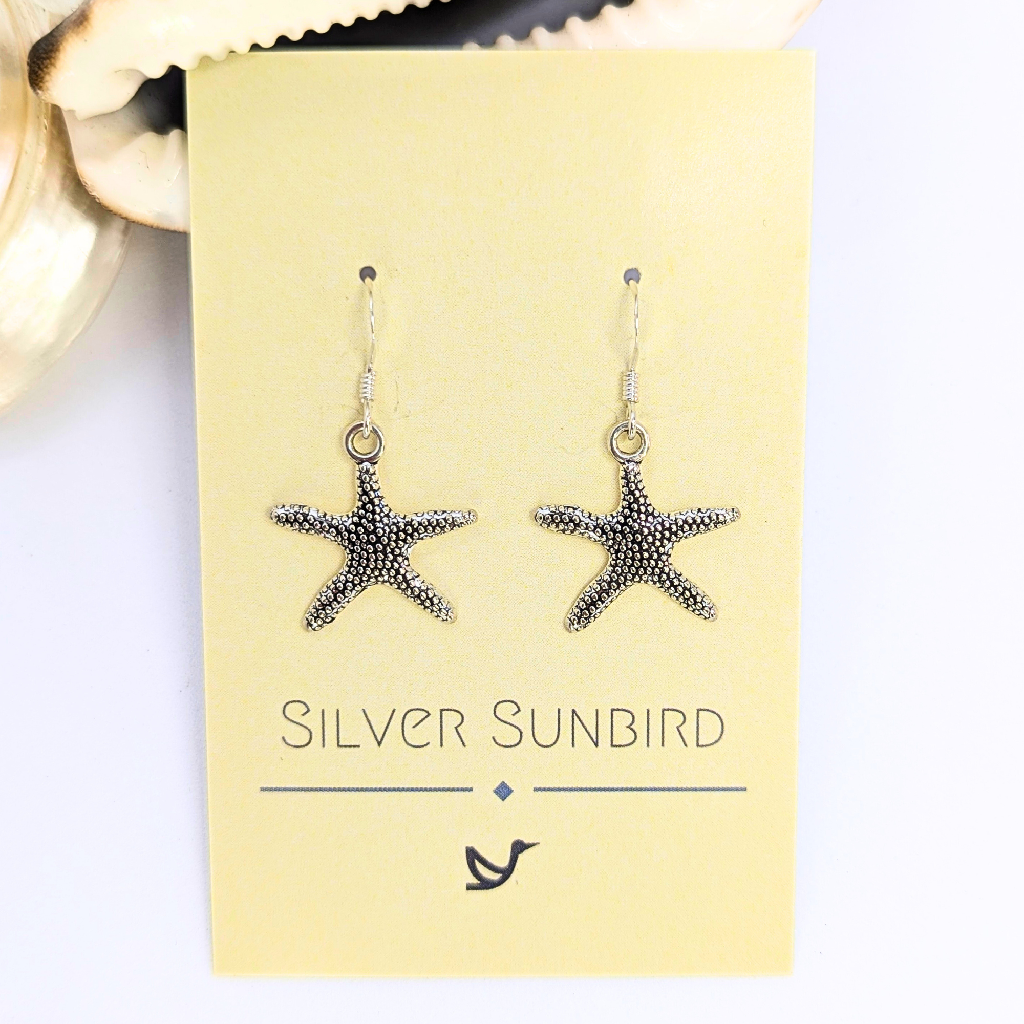 Detailed Silver Starfish Earrings by Silver Sunbird, shown on a pale yellow "Silver Sunbird" card with bird logo, set on a light background and seashell—ideal boho jewellery for ocean lovers.