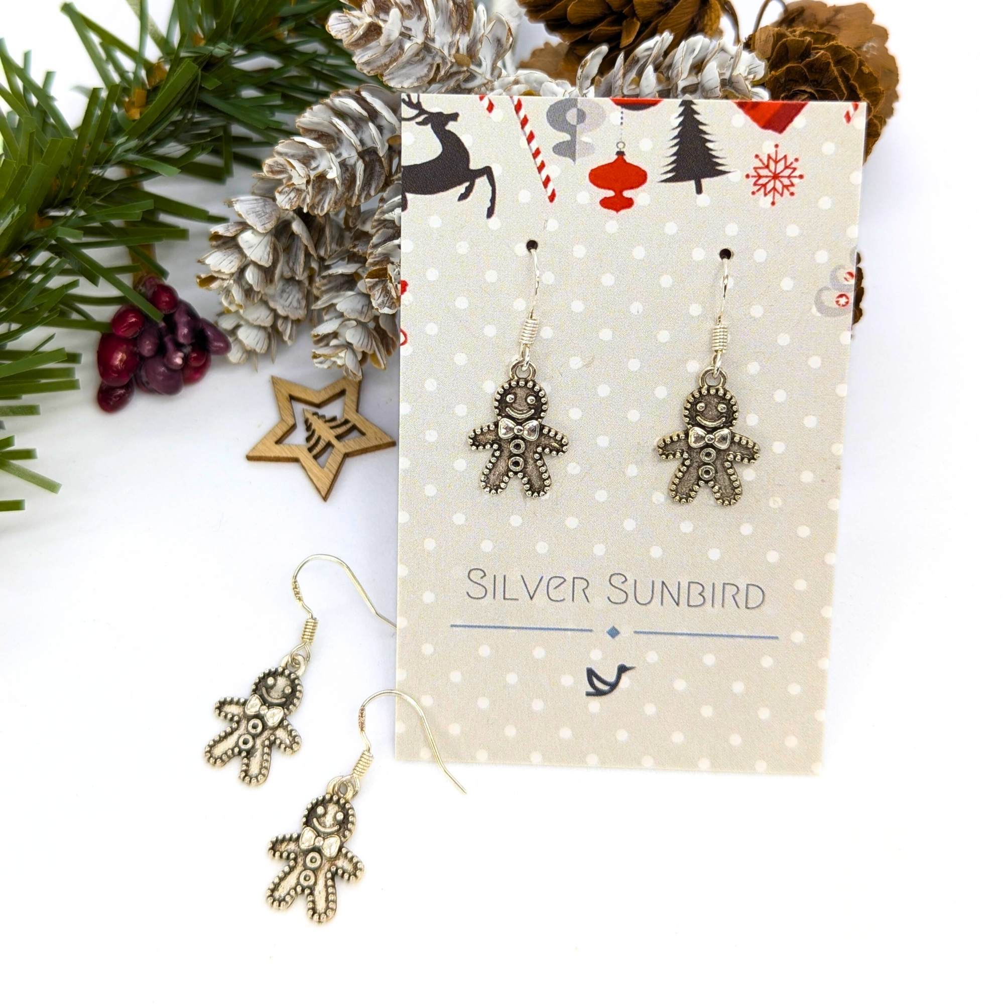 A festive earring display shows two pairs of Jolly Gingerbread Men Earrings by Silver Sunbird—crafted from 925 sterling silver—with one pair on a branded card and another pair in front, surrounded by pinecones and holiday jewelry décor.