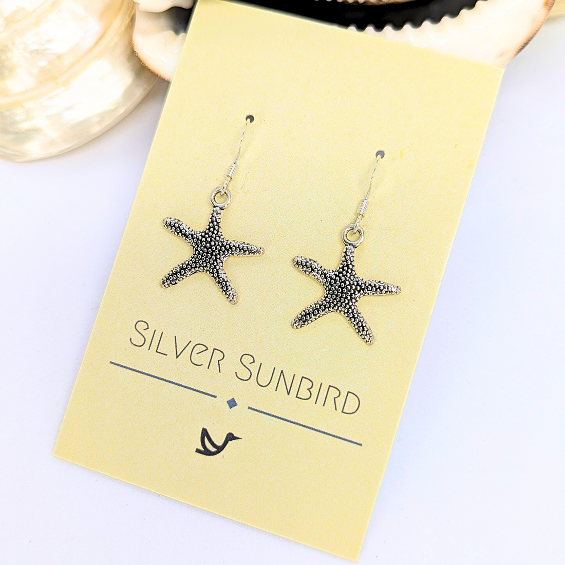 Detailed Silver Starfish Earrings by Silver Sunbird, displayed on a yellow branded card with a seashell accent in the background, are perfect for adding boho charm to any outfit.