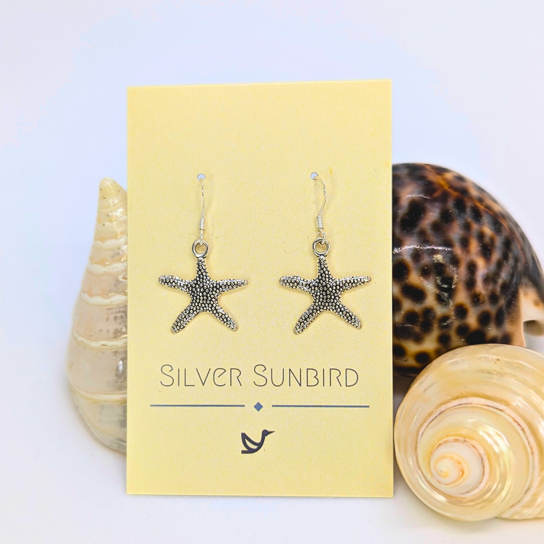 Detailed Silver Starfish Earrings by Silver Sunbird are showcased on a yellow card with three seashells in the background—an ideal gift for any boho jewellery lover.
