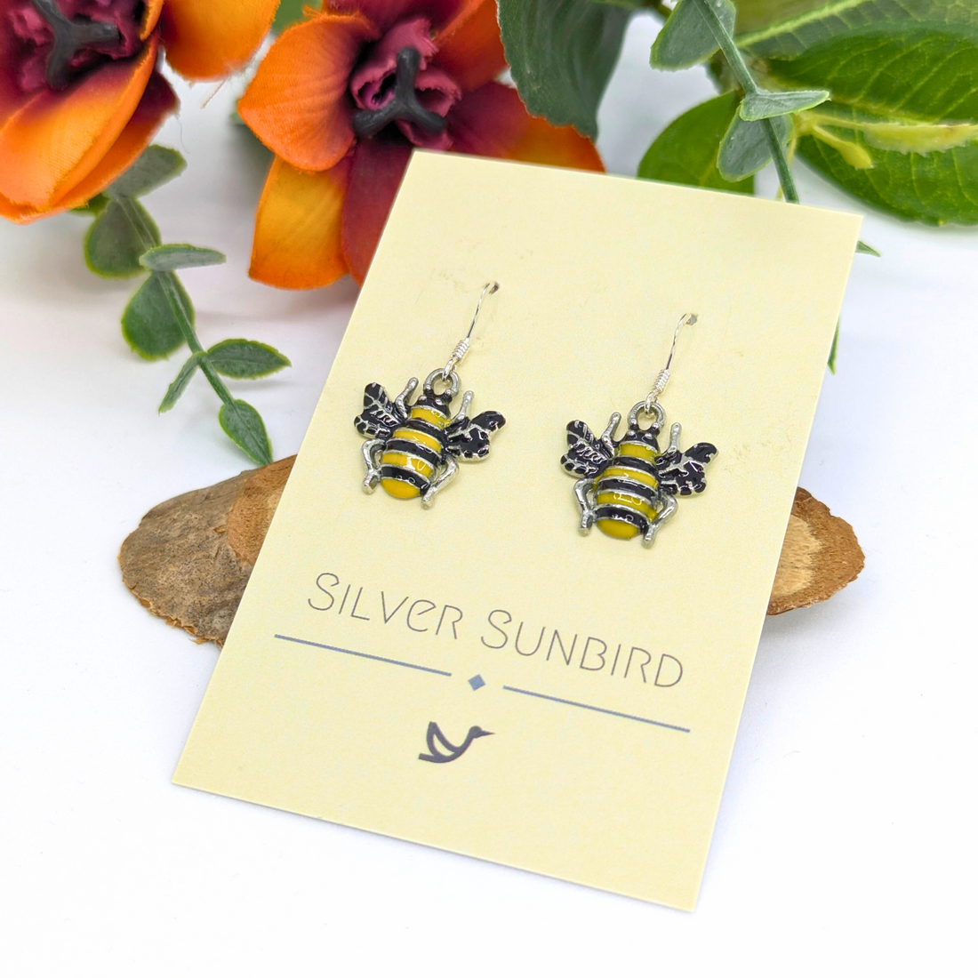 Bumble Bee Earrings by Silver Sunbird, featuring yellow and black stripes, displayed on a branded card amid orange flowers and green leaves.
