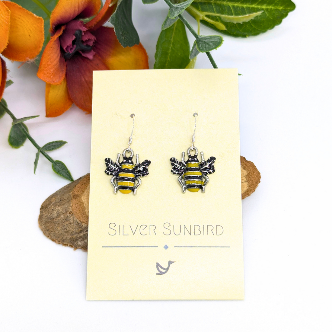 Bumble Bee Earrings by Silver Sunbird feature yellow and black enamel stripes and are displayed on a branded card, surrounded by artificial orange and green flowers.