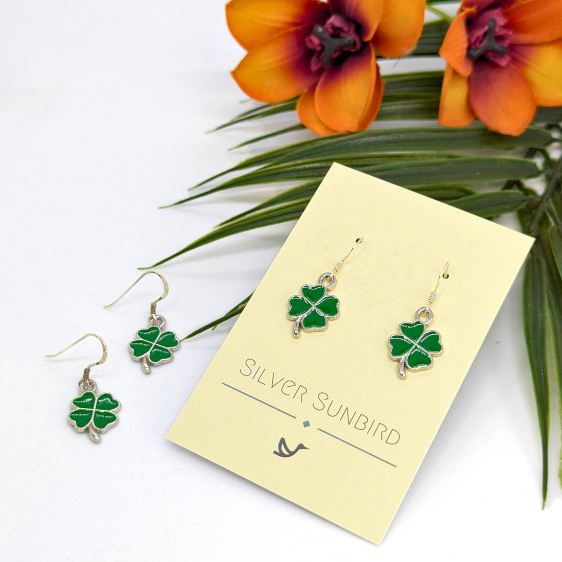 A pair of Lucky Four Leaf Clover Earrings by Silver Sunbird are shown on a branded card, with a second pair nearby. Above the handmade earrings, orange flowers and green leaves are arranged on a white background.