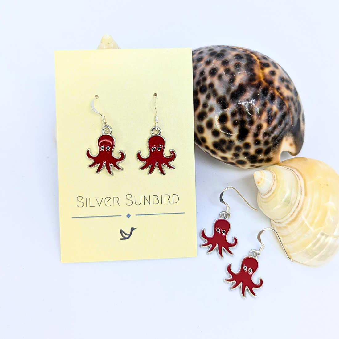 Two pairs of Silver Sunbird Red Octopus Earrings are shown, one on a branded card and one by seashells against a white background—a striking pick for anyone who loves ocean-inspired jewelry.