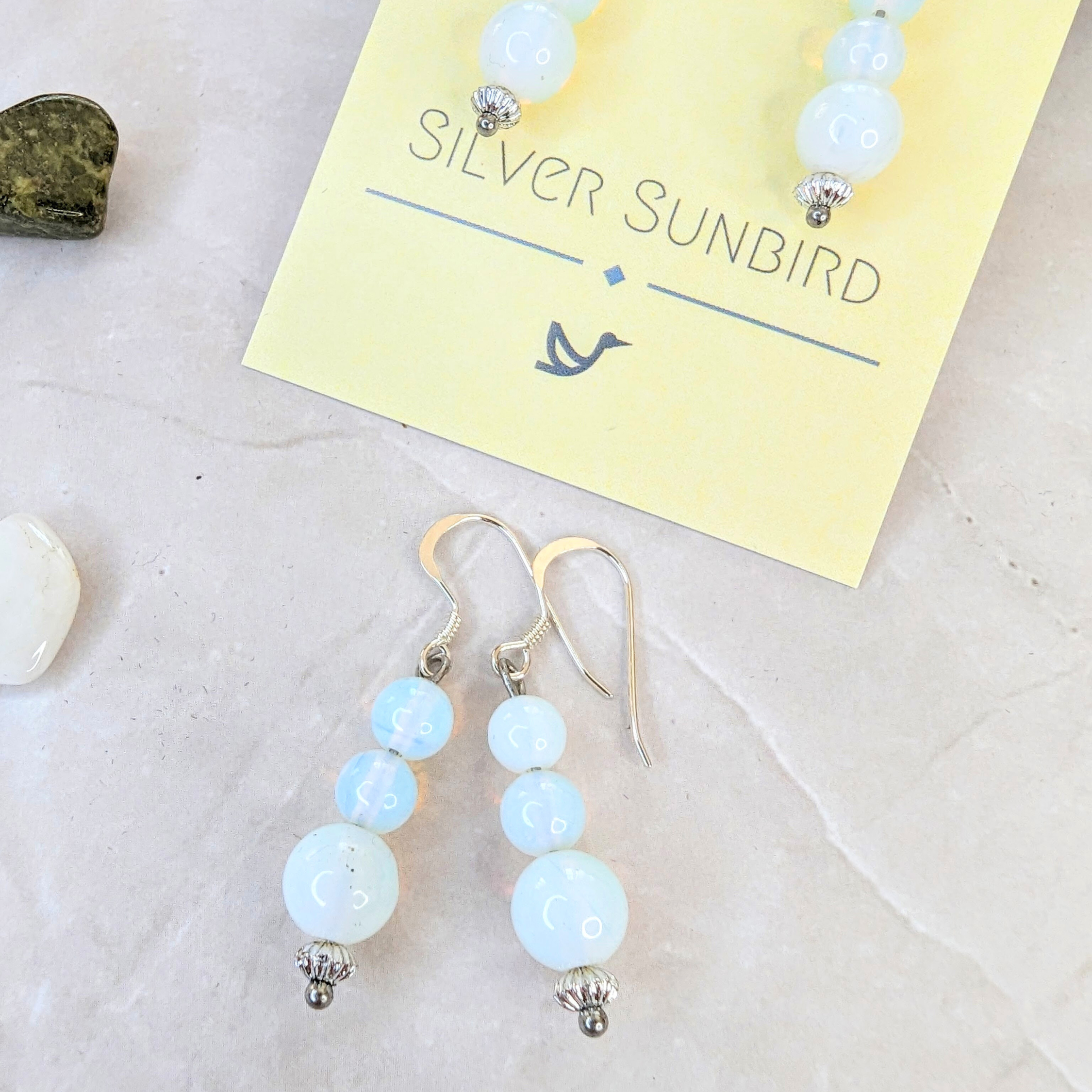 Opal Dangle Earrings by Silver Sunbird, featuring light blue beads and silver accents, are displayed on a brand card. The backdrop includes a marble surface adorned with a small dark stone and a white stone fragment.