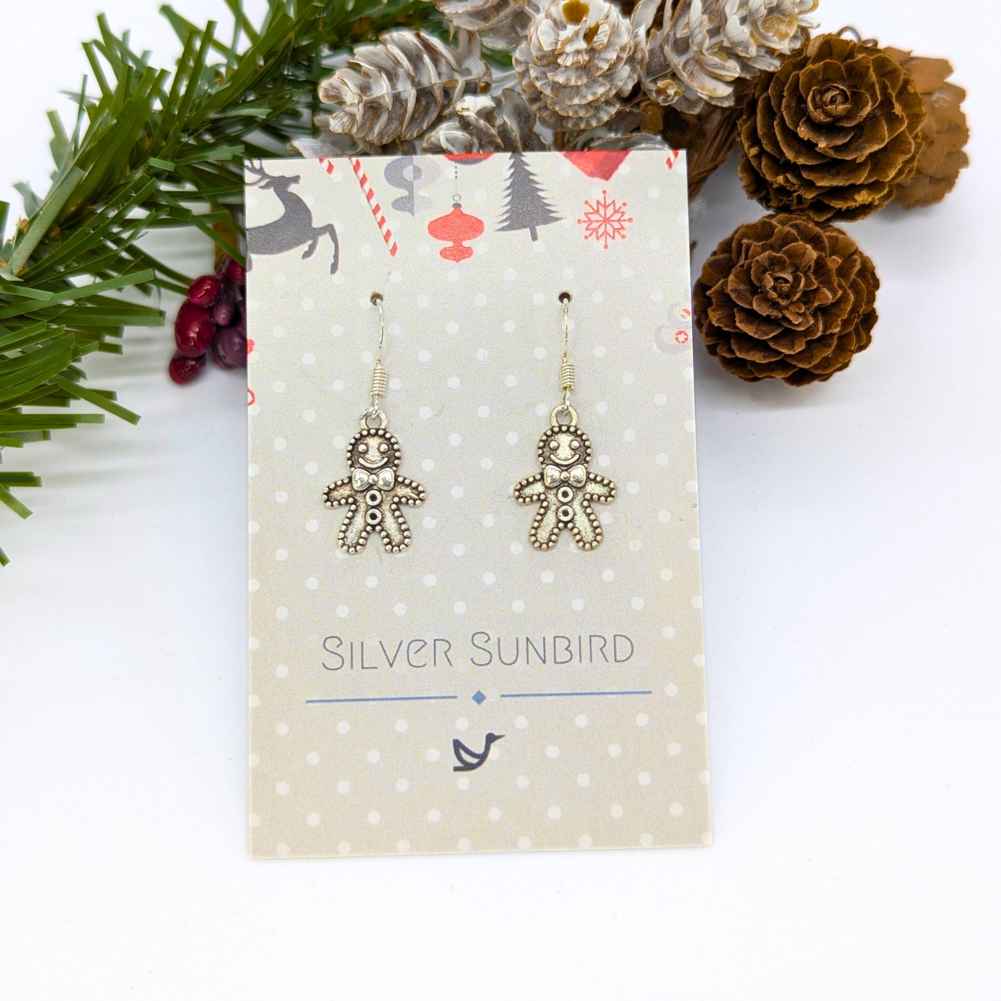 Jolly Gingerbread Men Earrings by Silver Sunbird, crafted in 925 sterling silver, come on a polka dot card—festive holiday jewelry displayed with pinecones, pine needles, and red berries, perfect for the season.