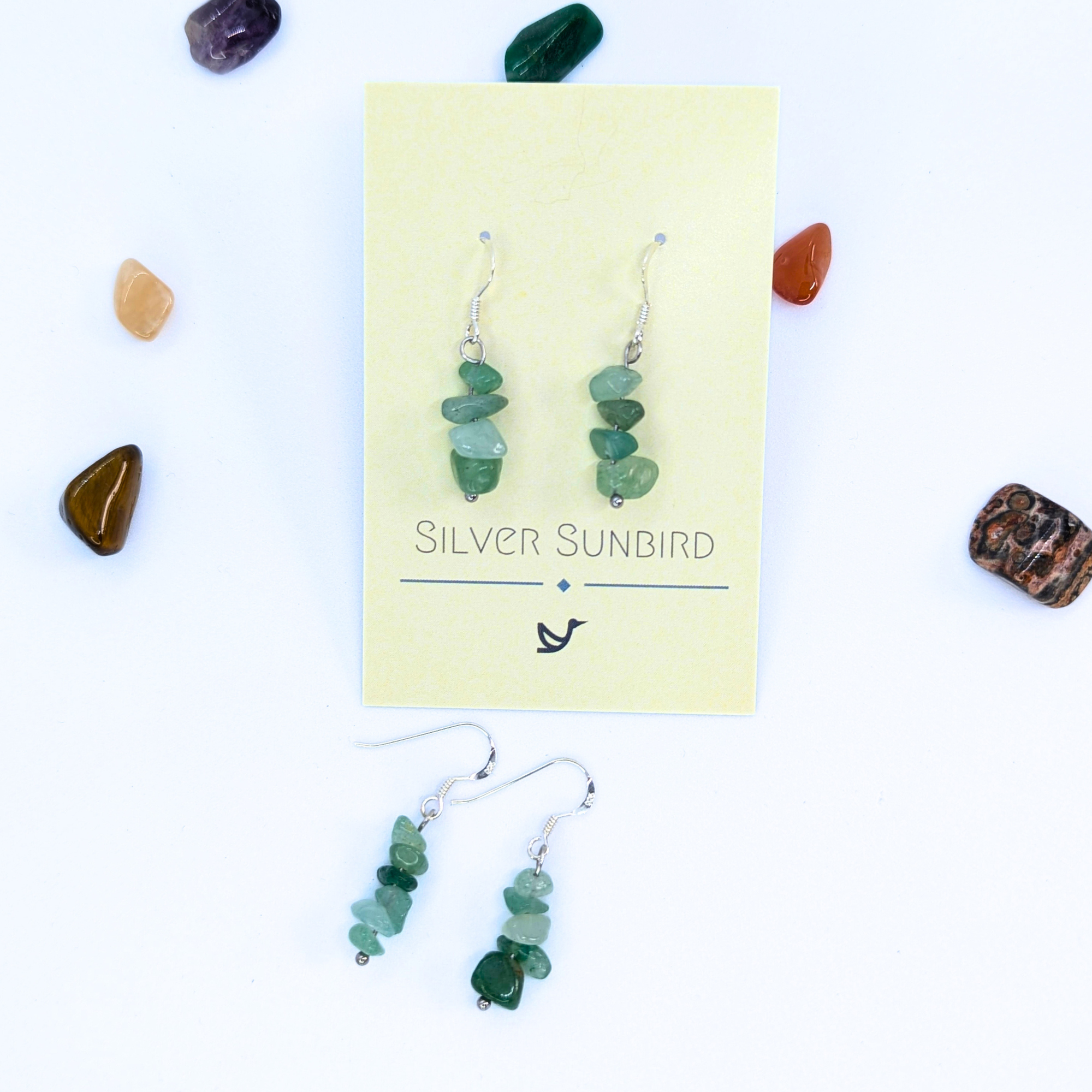 Green Aventurine Earrings by Silver Sunbird dangle from a branded card, surrounded by colorful polished stones—stunning handmade gemstone jewelry displayed on a white background.