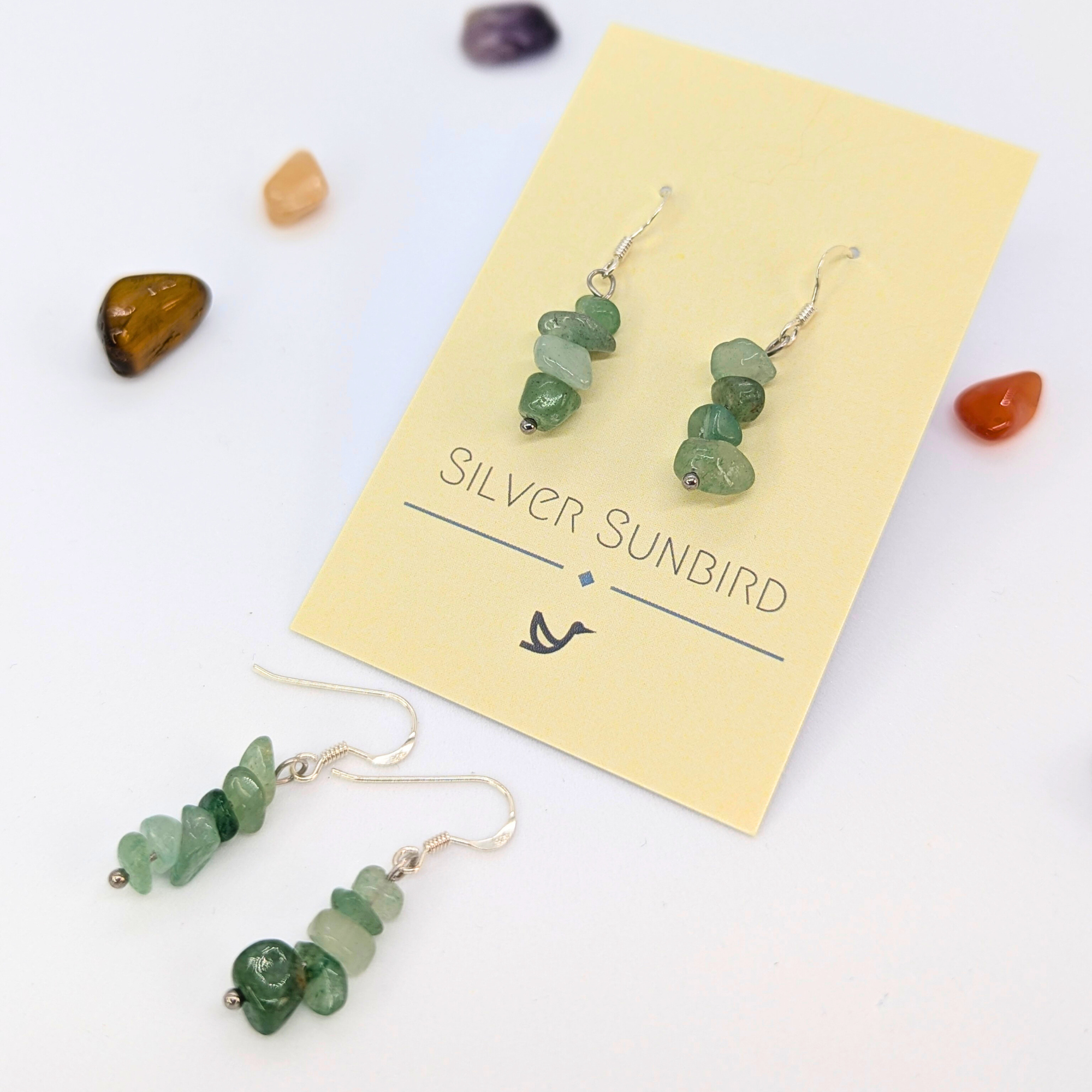 Green Aventurine Earrings by Silver Sunbird are displayed on a branded card, surrounded by colorful polished stones, highlighting elegant handmade gemstone jewelry.