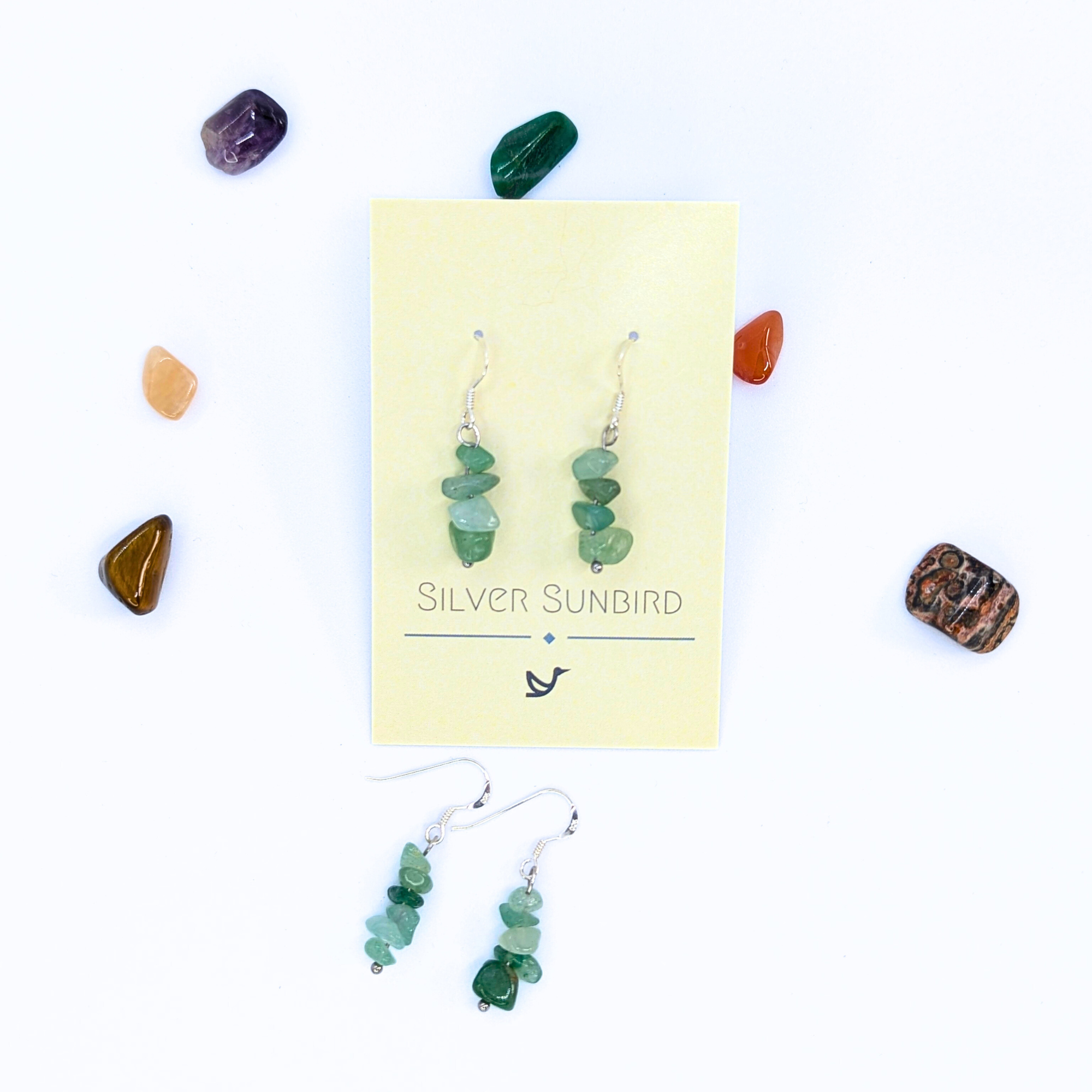 Green Aventurine Earrings by Silver Sunbird dangle from a branded card amid colorful polished stones, while another pair of sterling silver dangle earrings showcases the unique, handmade gemstone jewelry.