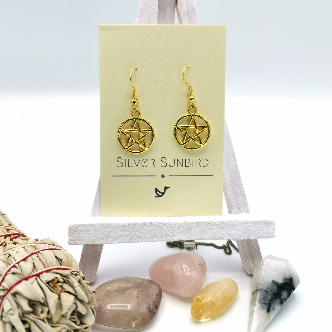 Pentagram Earrings by Silver Sunbird, rich in pagan symbolism, are displayed on a cream "Silver Sunbird" card atop a small white easel, with polished crystals and a red-tied sage bundle artfully arranged in front.