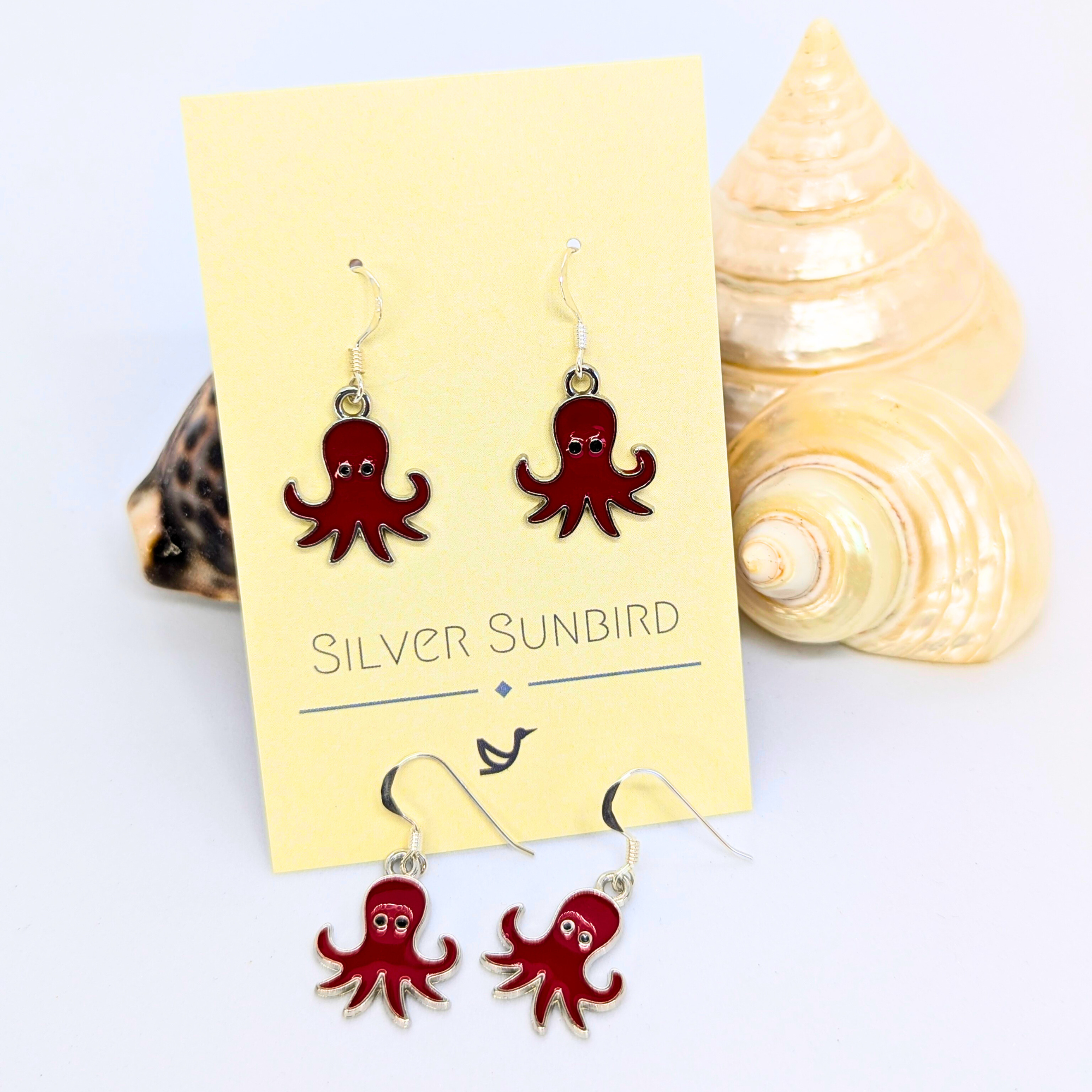 A pair of Silver Sunbird Red Octopus Earrings displayed on a branded card, with matching loose earrings nearby and cream and brown seashells accenting the ocean-inspired jewelry on a white surface.