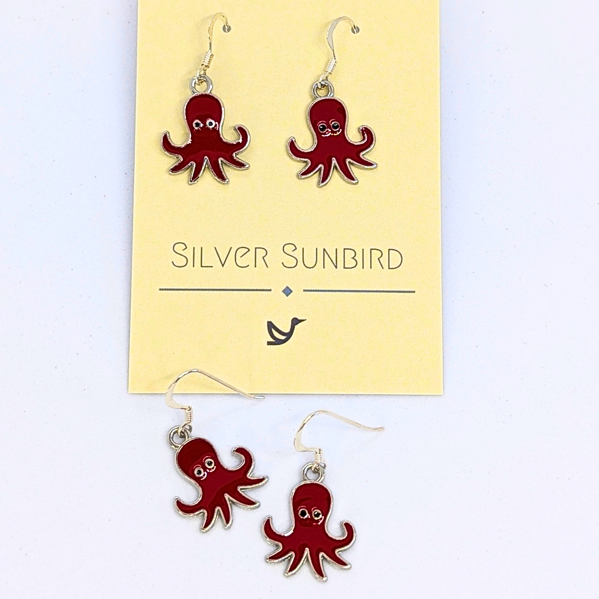 Red Octopus Earrings by Silver Sunbird feature an enamel finish and silver hooks, designed as ocean-inspired jewelry. Displayed on and below a branded "Silver Sunbird" card.