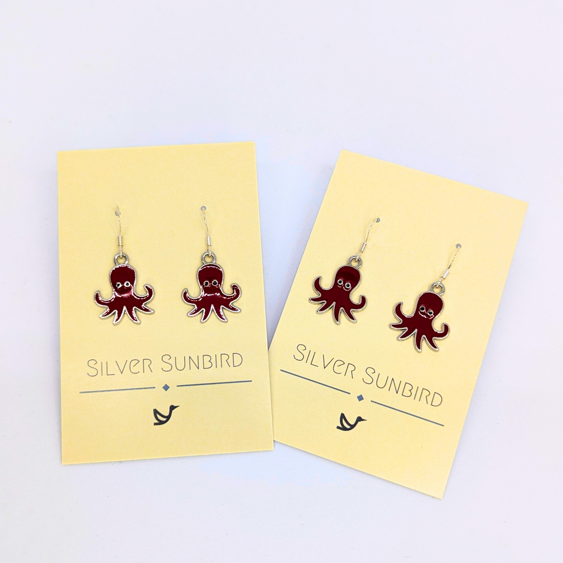 Two pairs of Red Octopus Earrings by Silver Sunbird hang on cream-colored branded cards, highlighting playful ocean-themed jewelry against a plain white backdrop.