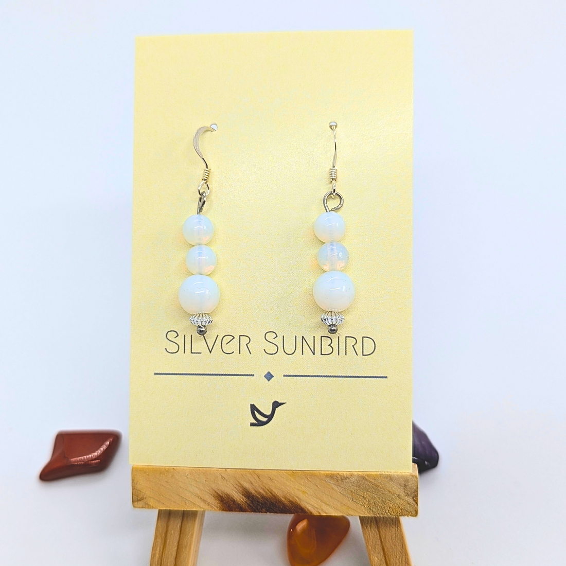 A pair of Opal Dangle Earrings by Silver Sunbird, featuring three pearl-like beads each, is displayed on a card labeled "Silver Sunbird," placed on a small wooden easel with decorative stones in the background.