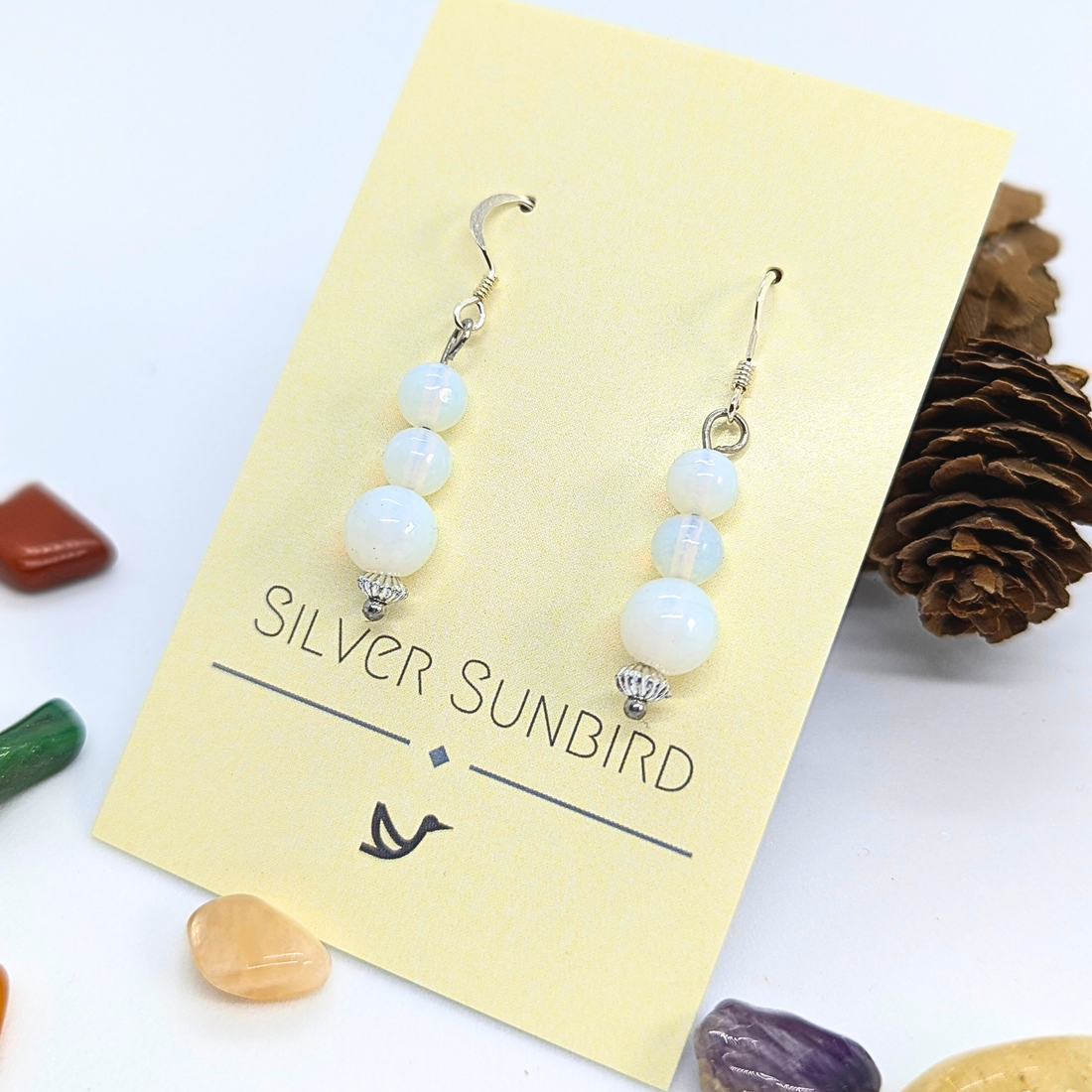A pair of Opal Dangle Earrings by Silver Sunbird, featuring three white round beads on each earring, displayed on a yellow card with colorful stones and a pine cone.