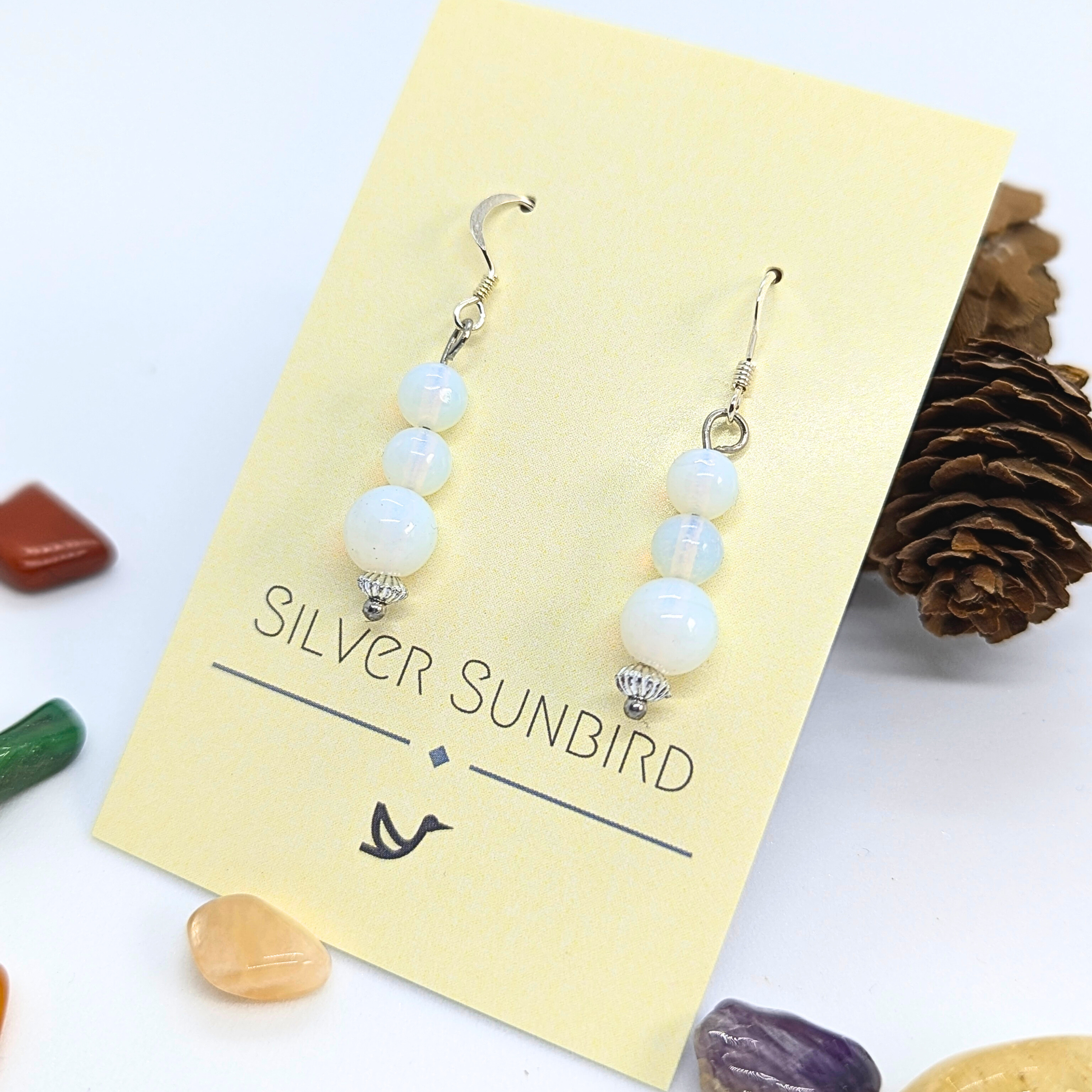 A pair of Opal Dangle Earrings by Silver Sunbird, featuring three white round beads on each earring, displayed on a yellow card with colorful stones and a pine cone.