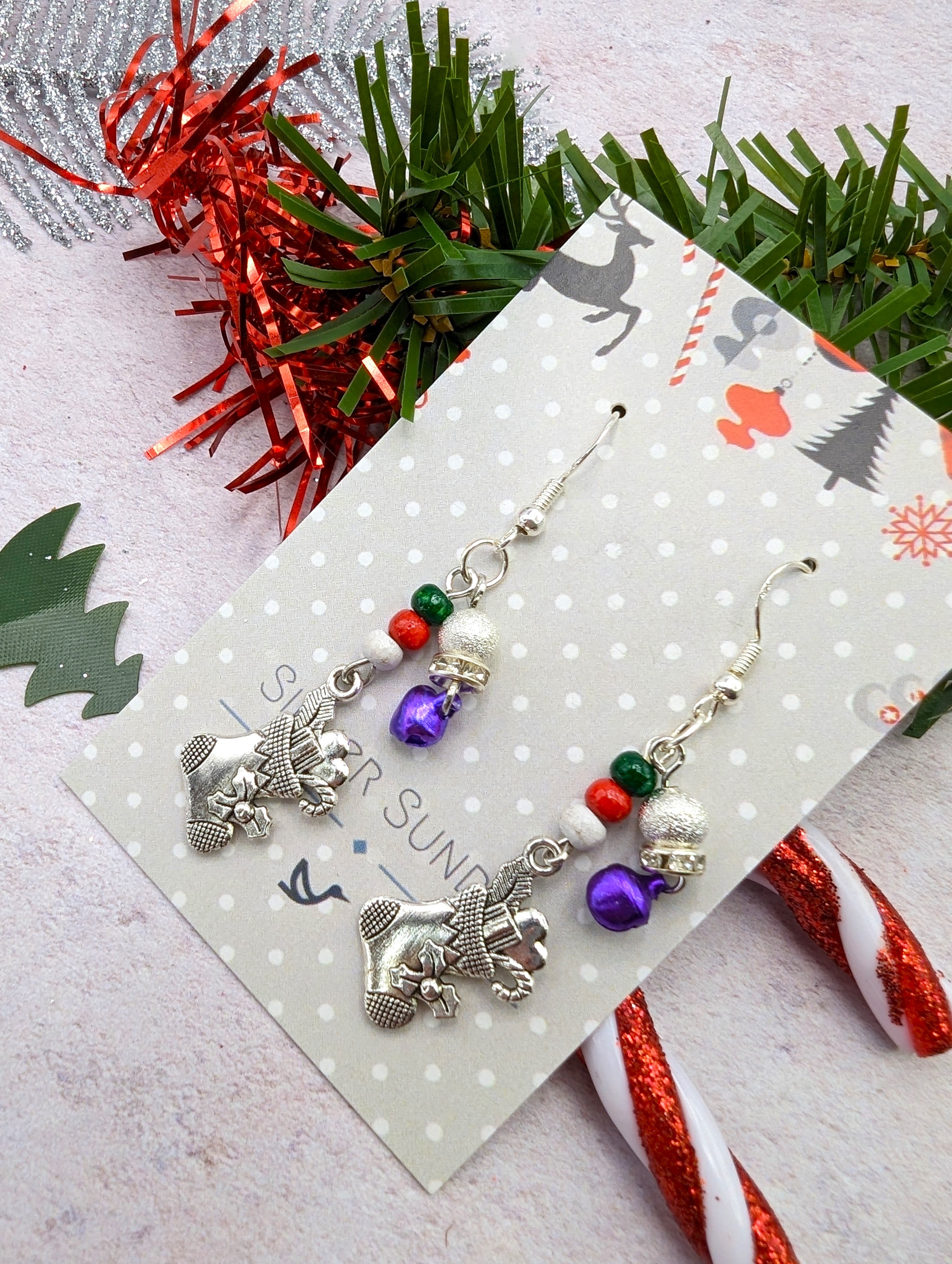 The Jingle Stockings Earrings by Silver Sunbird showcase sterling silver stocking charms, red and green beads, and purple baubles presented on a decorative card. The festive presentation is further adorned with candy canes and green tinsel highlighted in red.