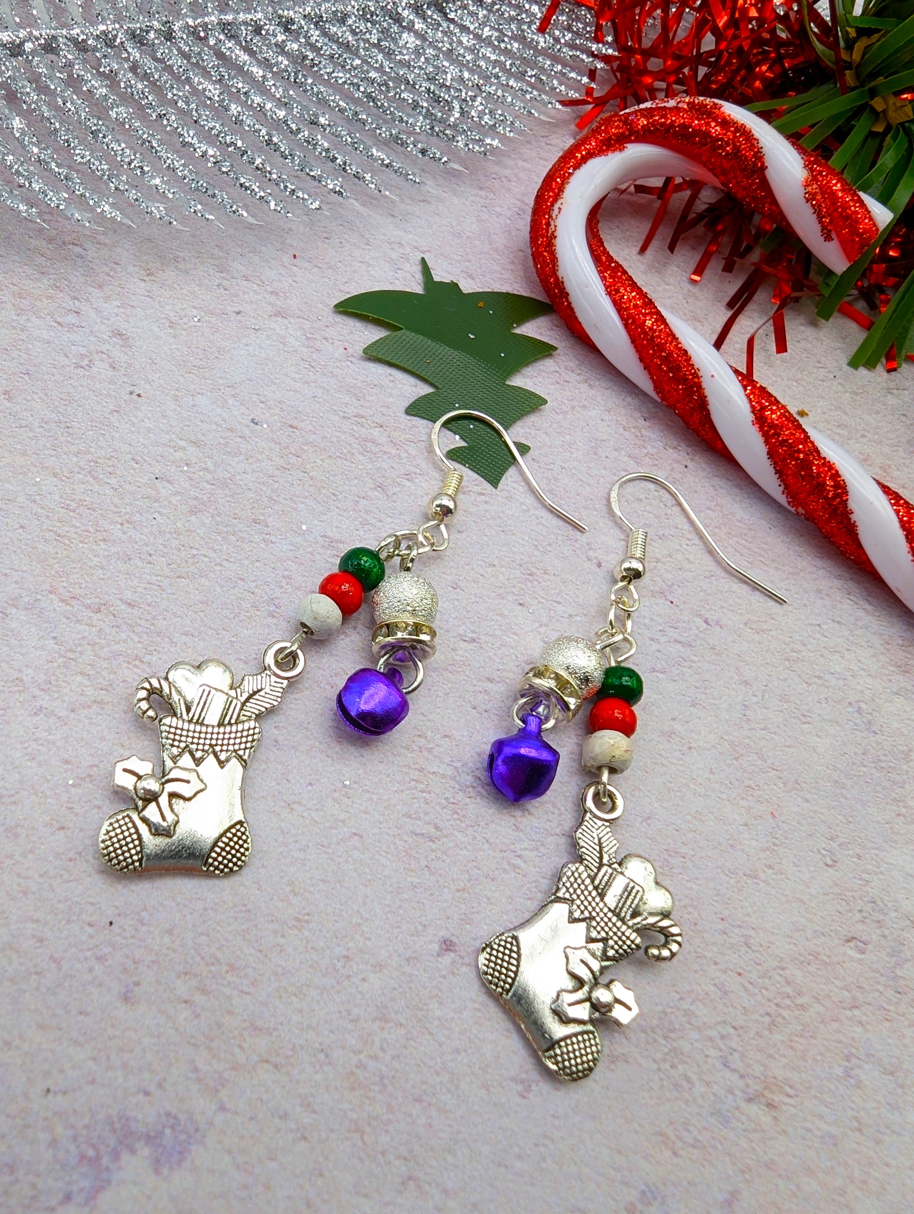 Introducing the Jingle Stockings Earrings by Silver Sunbird, featuring festive handmade Christmas designs crafted with sterling silver stockings, colorful beads, purple and silver bells, and green tree-shaped decorations. The background is beautifully accented with a candy cane, silver ribbon, and red tinsel on a textured surface.