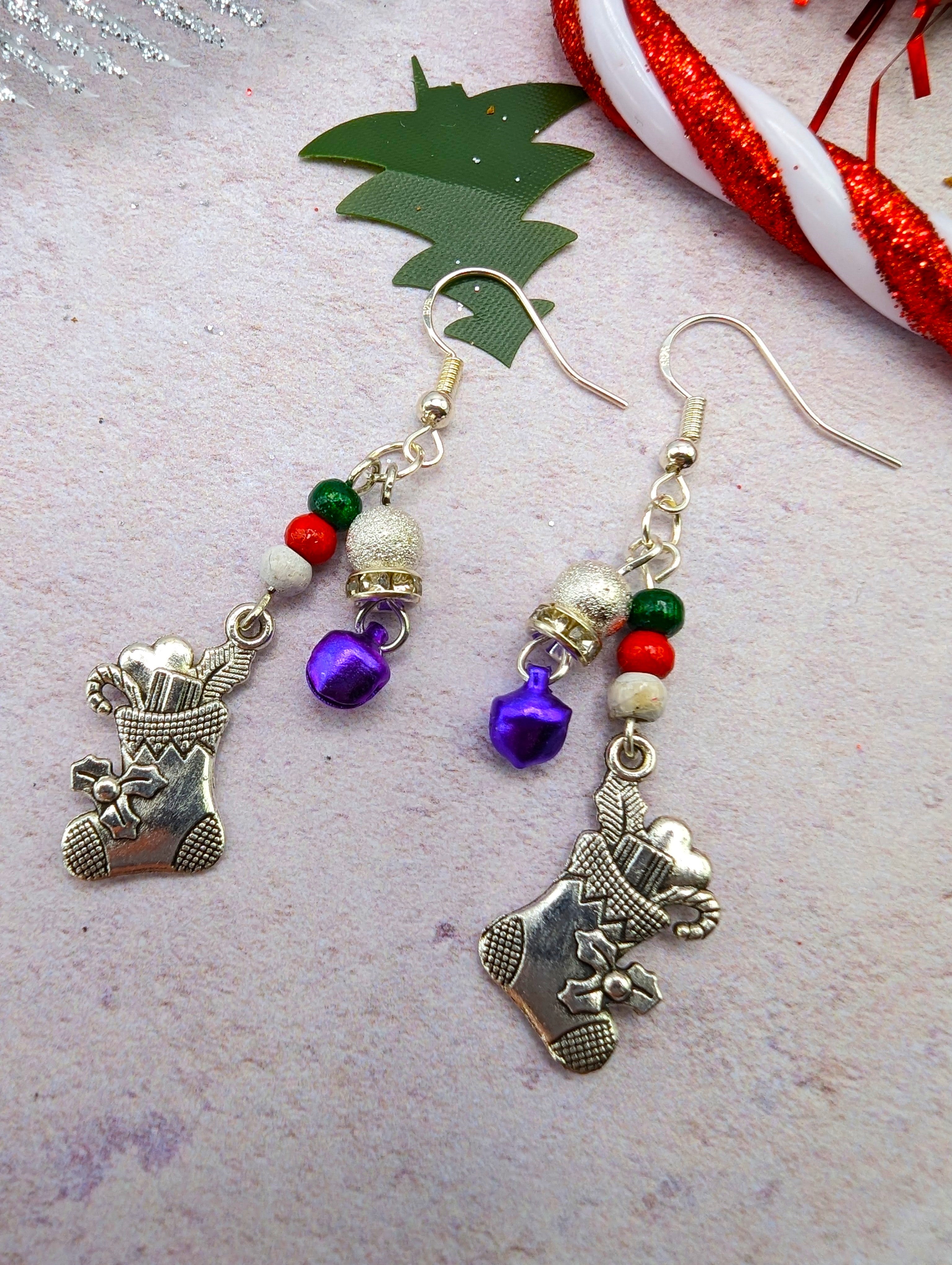 Introducing the Jingle Stockings Earrings by Silver Sunbird: handcrafted festive jewelry with charming silver stockings, vibrant red and green beads, whimsical purple bells, and elegant white accents. These sterling silver earrings are showcased on a light backdrop adorned with decorative greenery and a candy cane ribbon, making them the perfect holiday accessory.