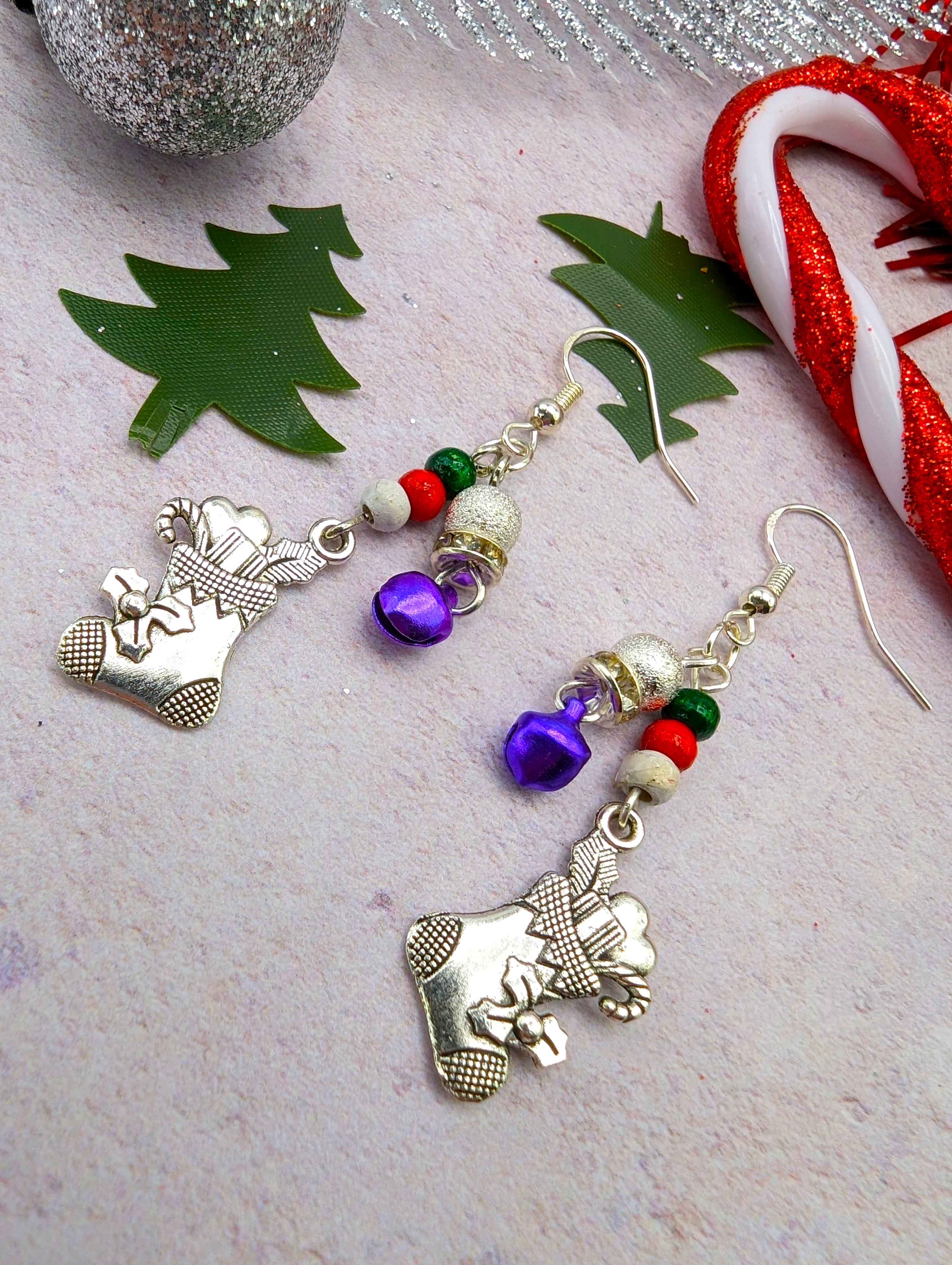 Introducing the Jingle Stockings Earrings by Silver Sunbird, festive handmade jewelry featuring sterling silver stocking charms adorned with purple, red, and green beads. Set against a backdrop of candy canes and tree cutouts, these earrings beautifully capture the holiday spirit.