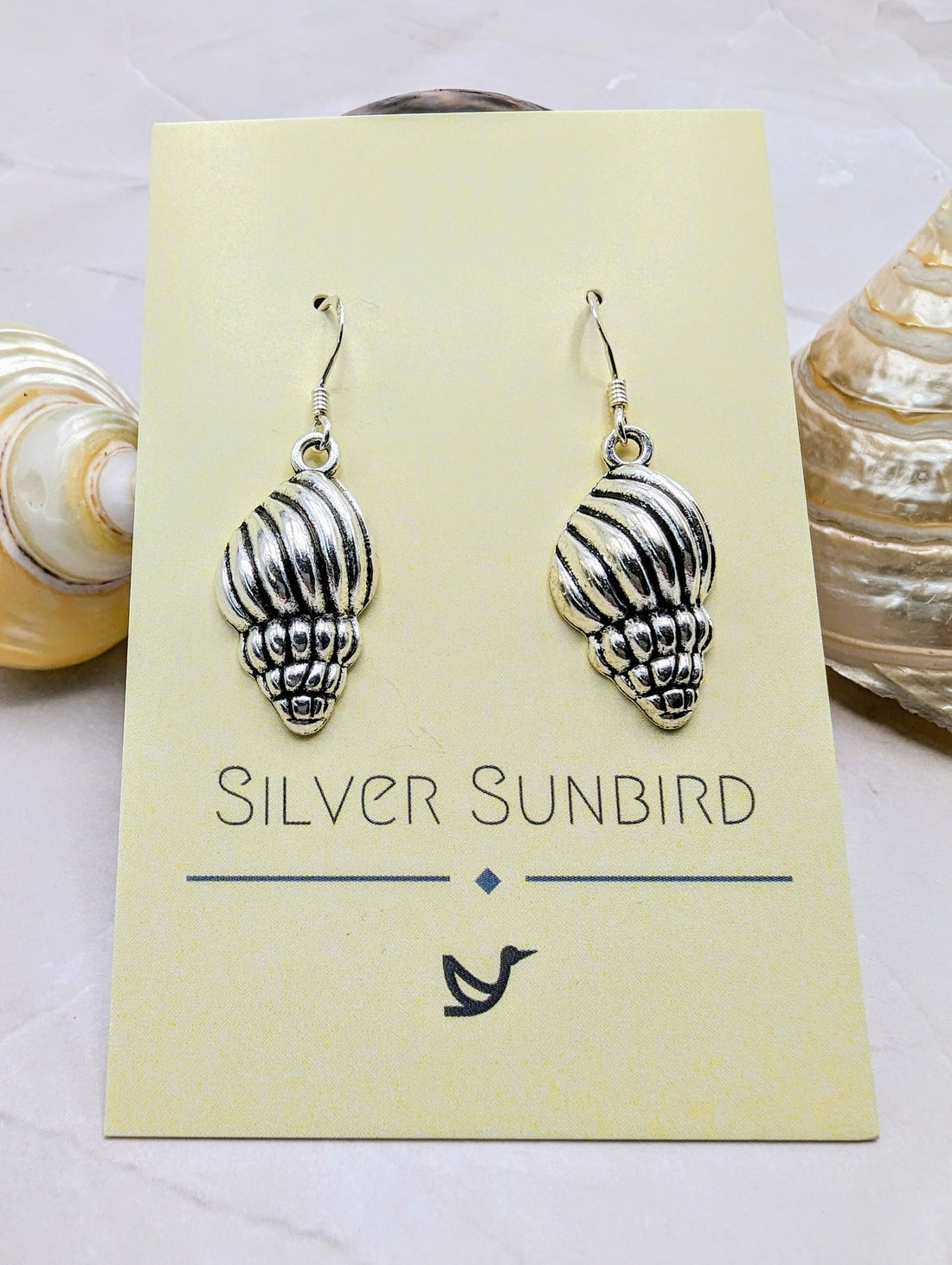 Two Cool Conch Earrings by Silver Sunbird, elegantly shaped like seashells, are displayed on a yellow card. The background includes seashells on a textured surface. These hypoallergenic earrings combine elegance and comfort.