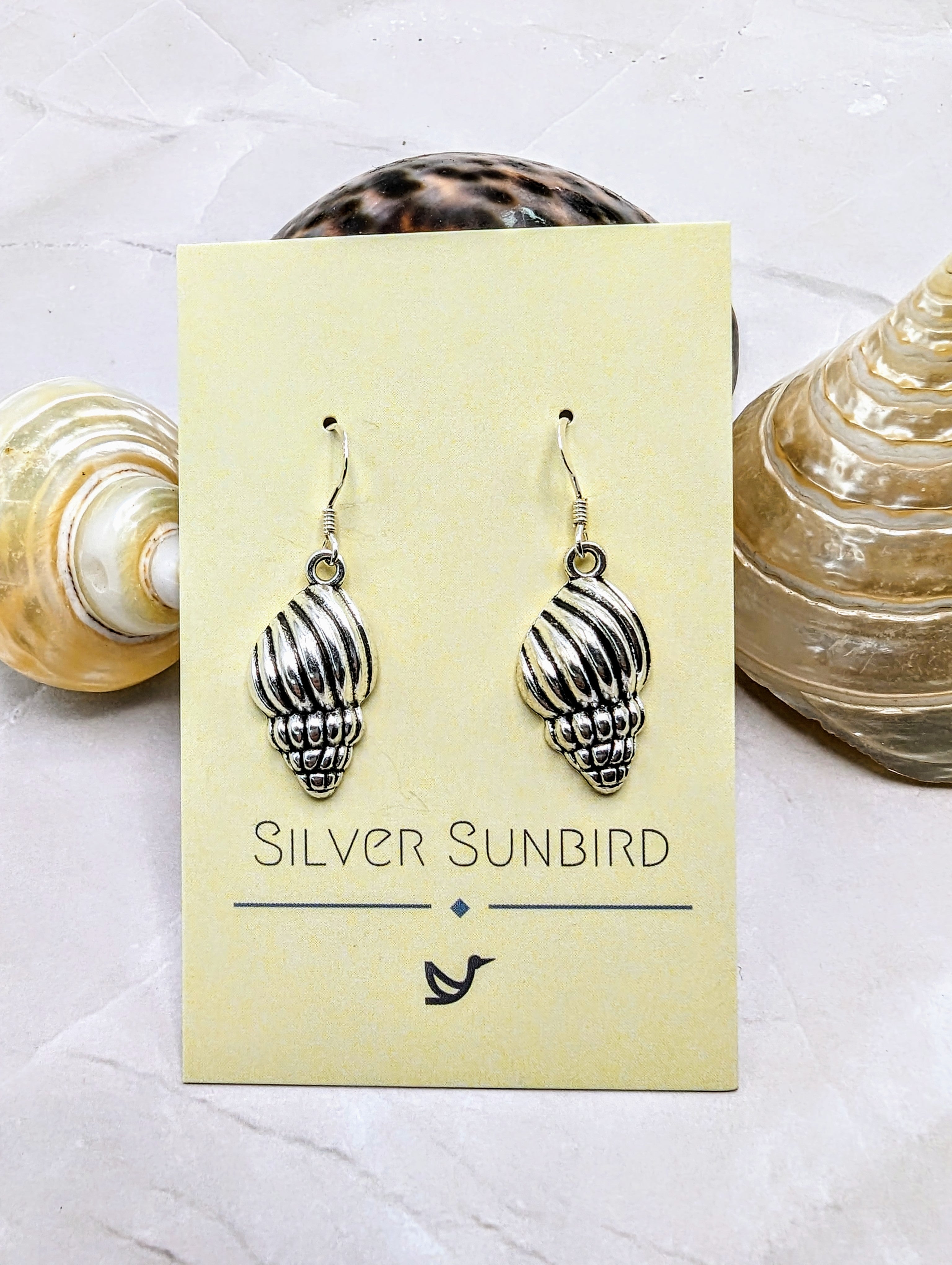 A close-up of Cool Conch Earrings by Silver Sunbird shows them hanging on a branded card, placed on a marble surface surrounded by seashells, capturing their essence.