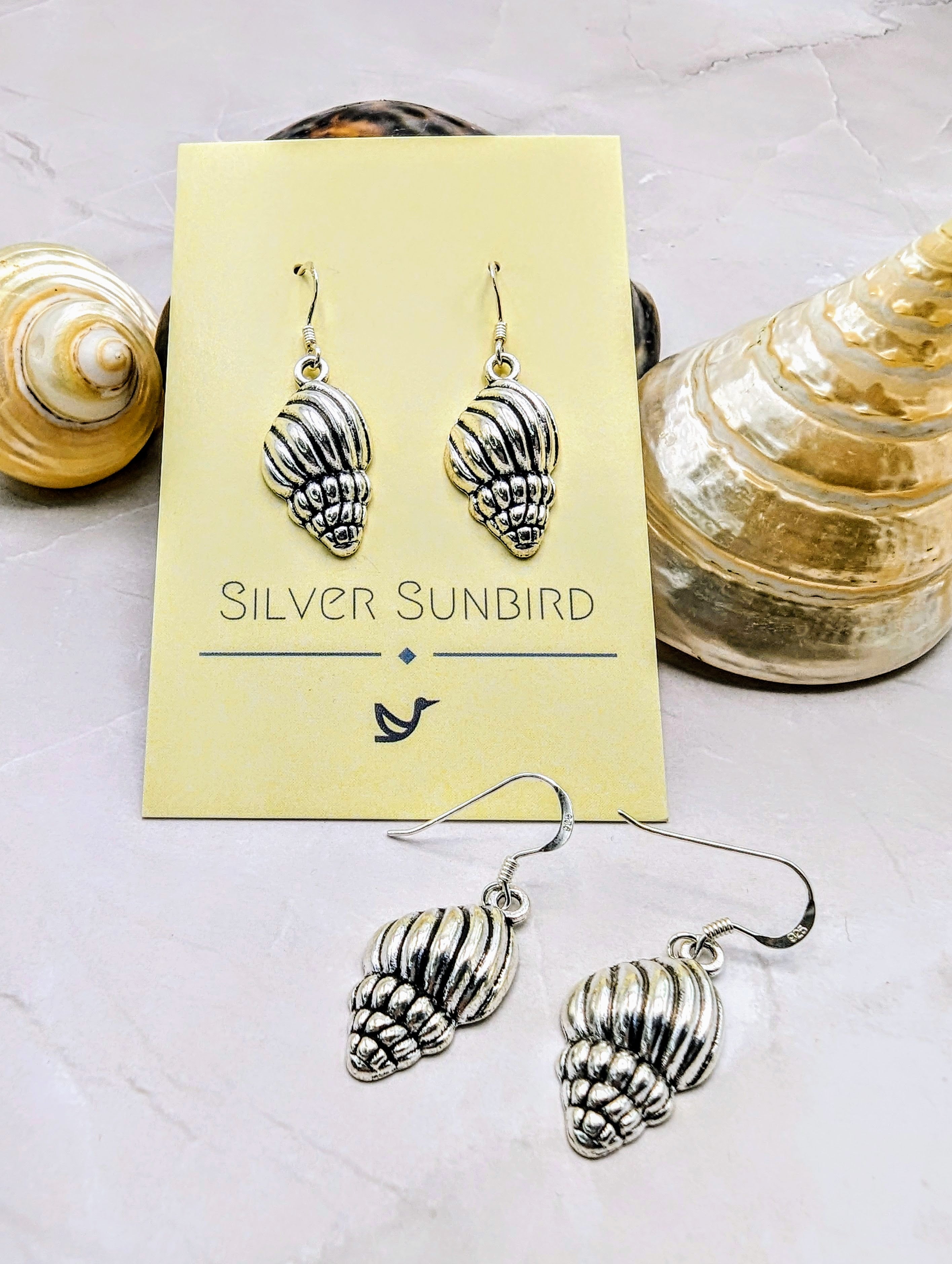 The Cool Conch Earrings, crafted from sterling silver and branded by Silver Sunbird, are elegantly presented on a card. Two earrings hang above with two below amidst scattered seashells, enhancing the coastal vibe. These hypoallergenic earrings offer all-day comfort.