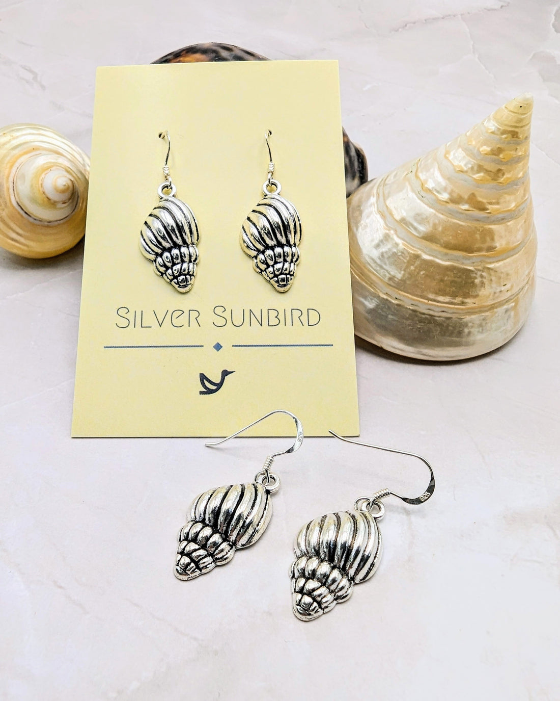 The Cool Conch Earrings, by Silver Sunbird, are elegantly displayed on their branded card atop a marble surface, with scattered seashells in the background to enhance the sea shell theme.