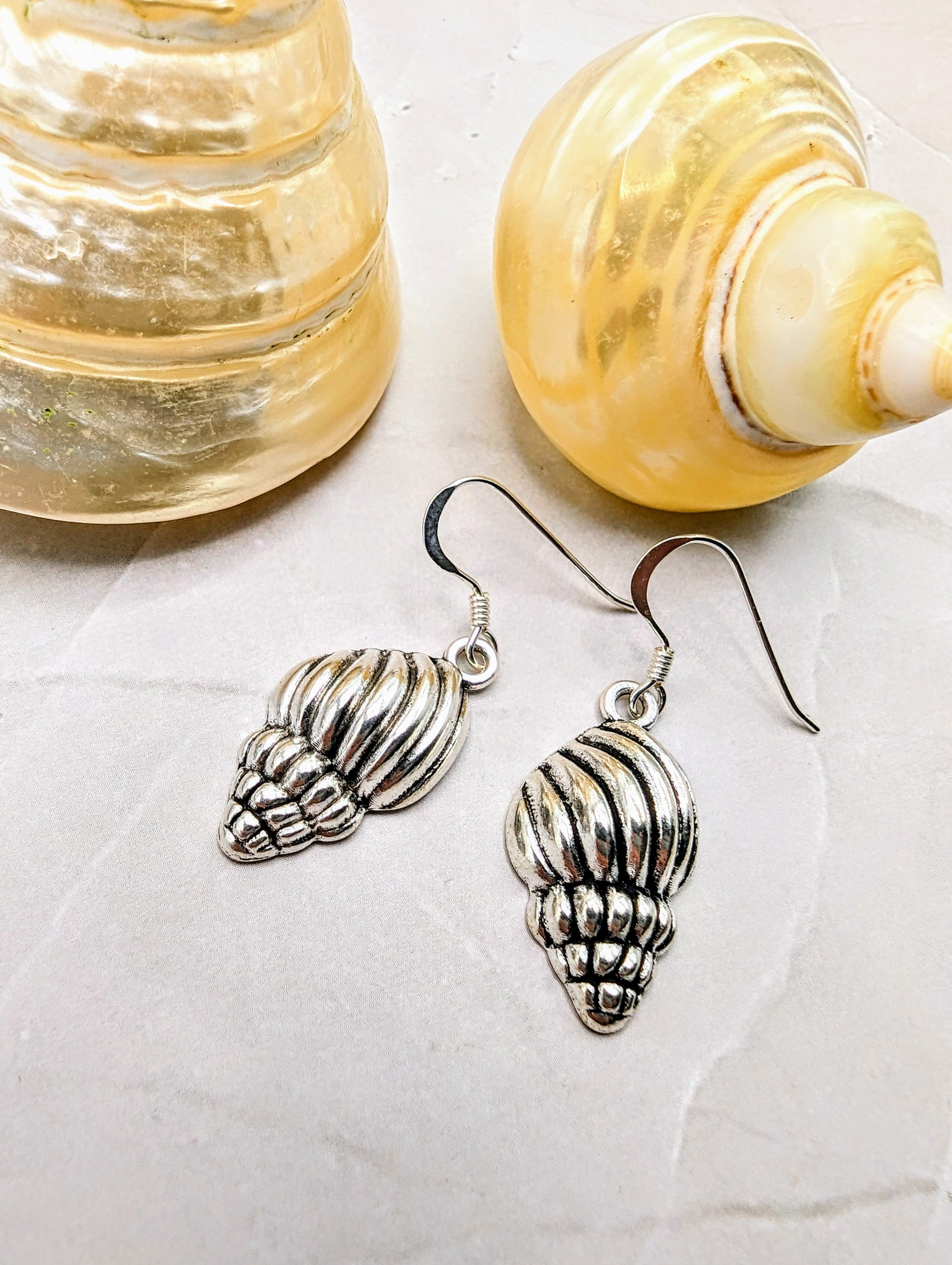 The Cool Conch Earrings by Silver Sunbird sit on a textured surface beside two shiny seashells.