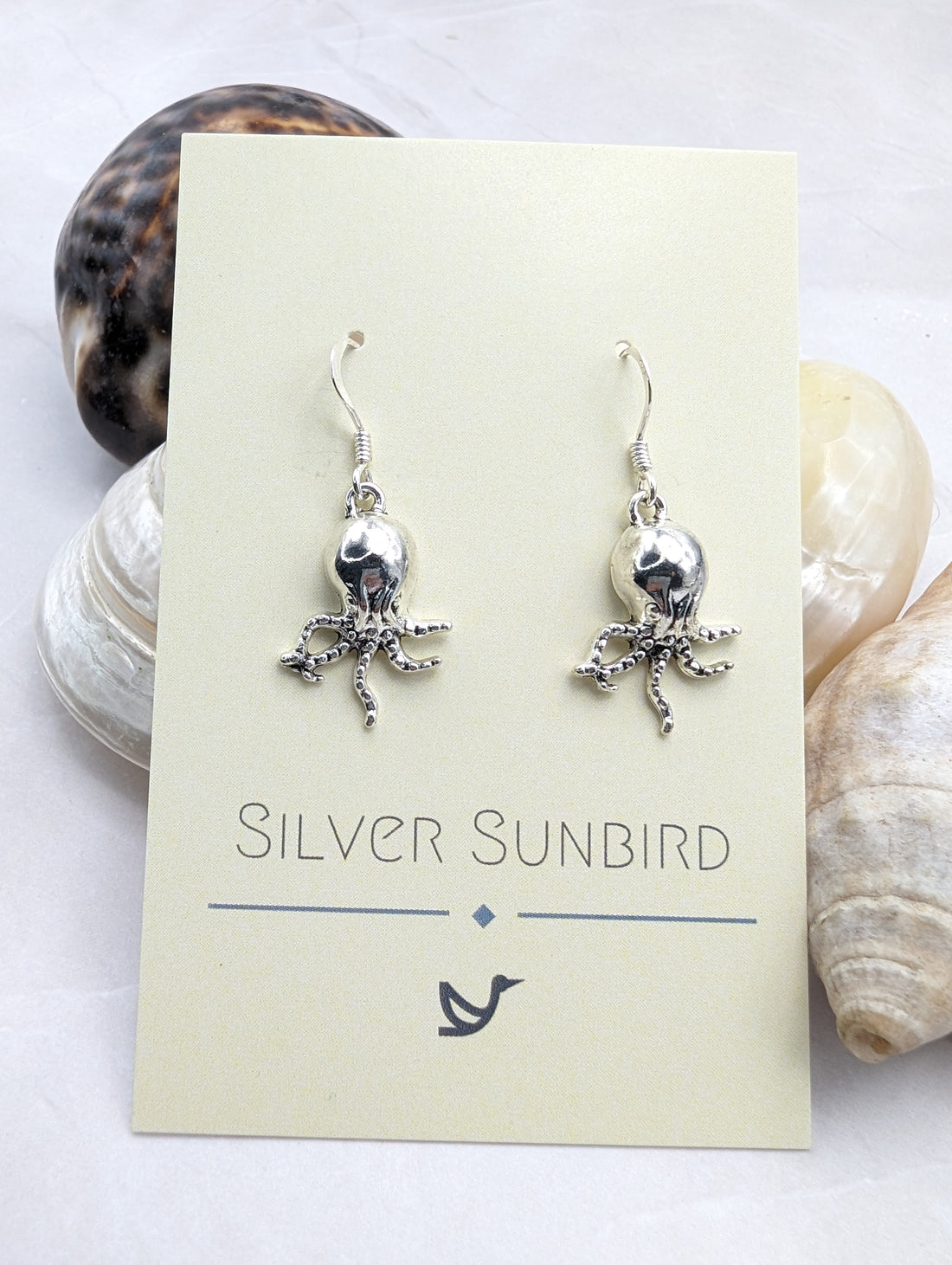 A card from "Silver Sunbird" showcases a pair of Awesome Octopus Earrings made from sterling silver, featuring intricate octopus charms. Displayed against an assortment of seashells on a white surface, the card highlights the brand name and includes a small bird logo at the bottom, adding an elegant touch to any ocean-inspired jewellery collection.