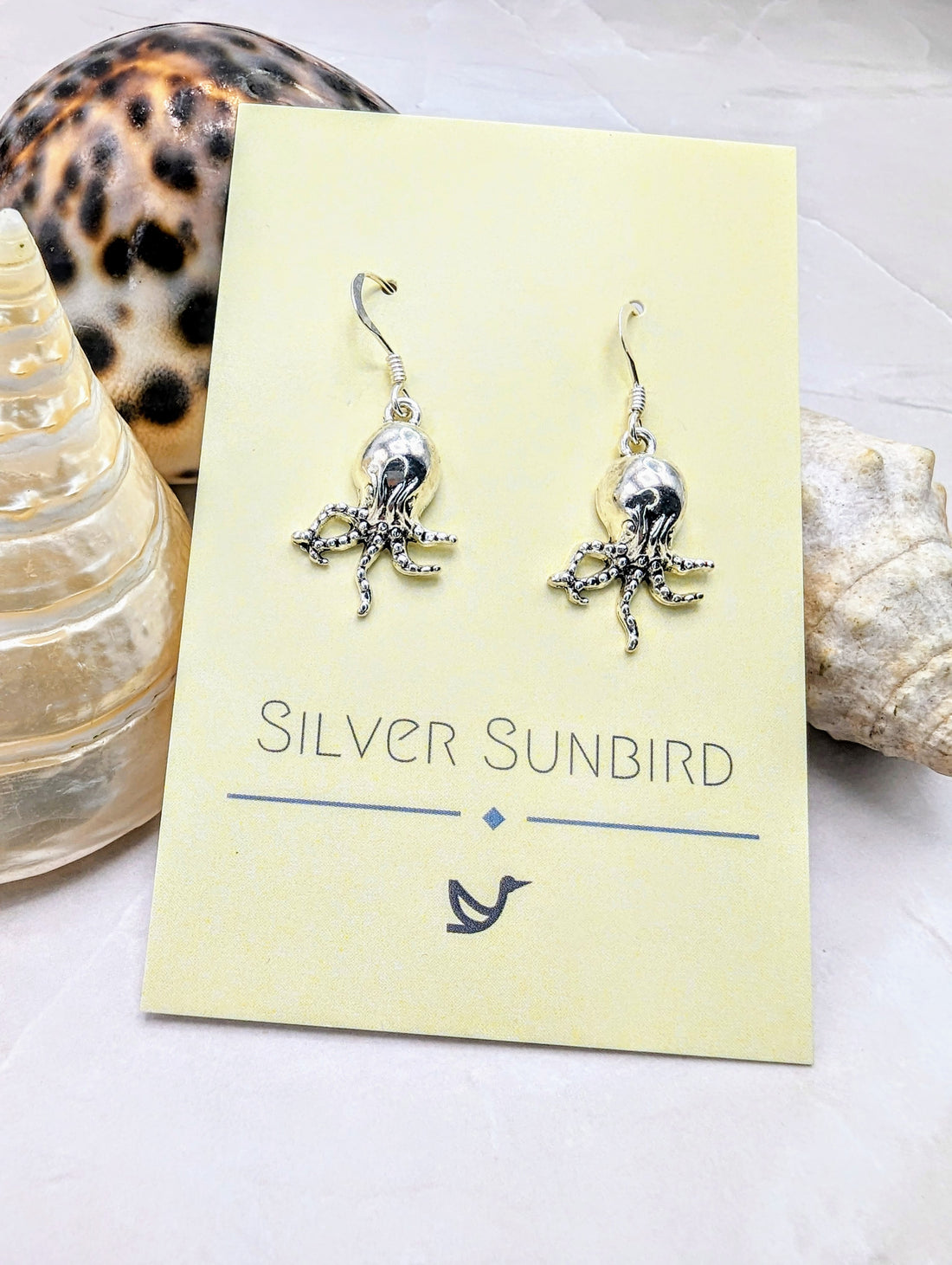 Awesome Octopus Earrings from Silver Sunbird are displayed on a yellow card against a light-coloured background. The display is set among three seashells, one conical and two varied in shape, capturing the essence of Ocean Jewellery.