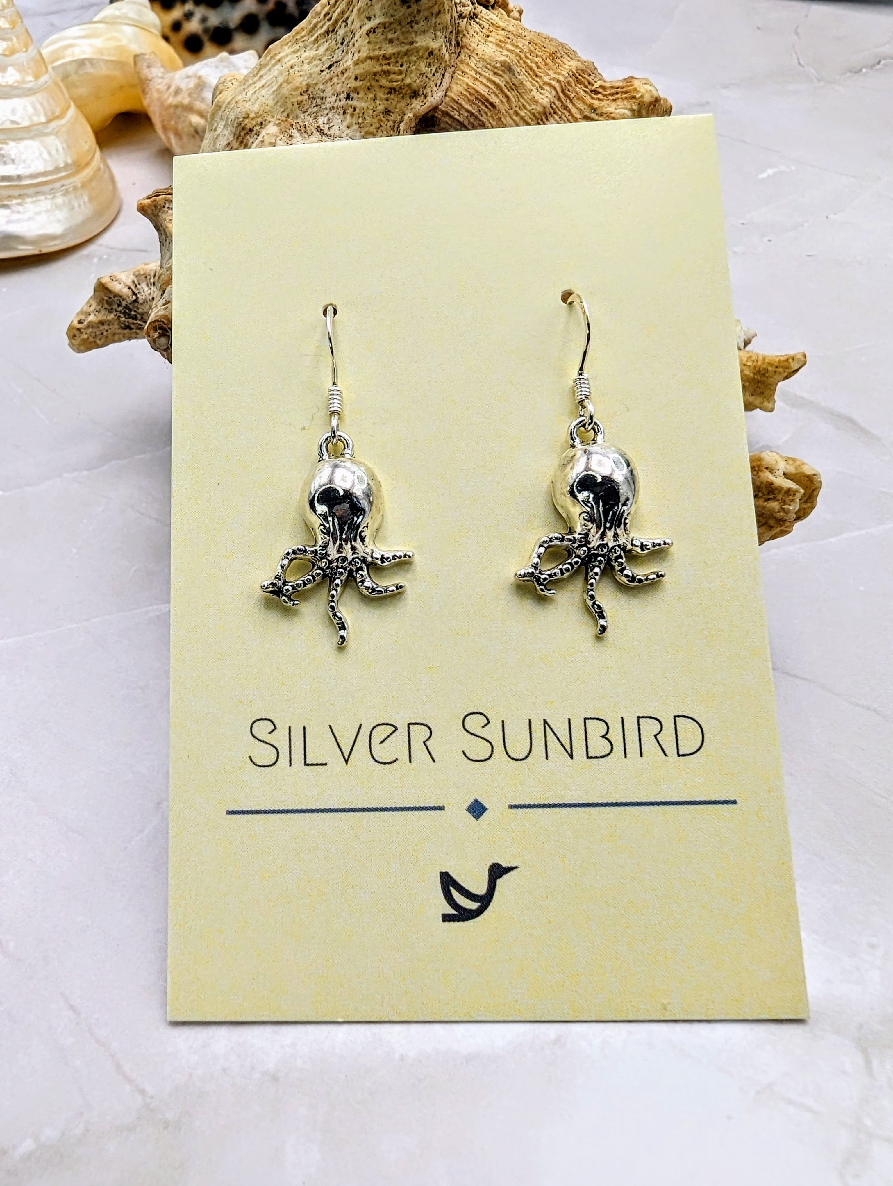 A display card labelled "Silver Sunbird" features dangling sterling silver "Awesome Octopus Earrings." The card rests on a light-coloured surface, with a piece of textured driftwood in the background, evoking the allure of ocean jewellery.
