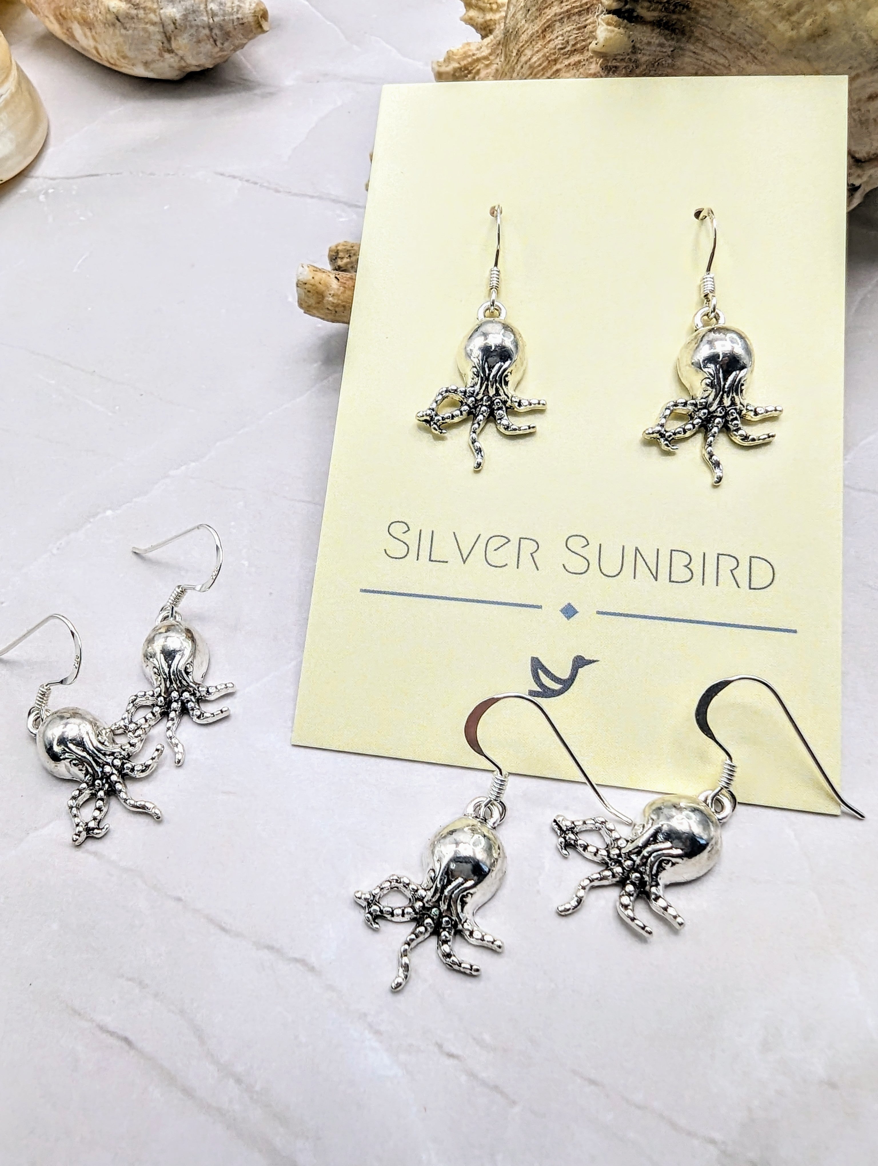 A display of Awesome Octopus Earrings on a light-coloured surface. Two pairs of earrings are shown, one on a card labelled "Silver Sunbird" and another pair lying flat nearby. The ocean jewellery features detailed octopus designs with dangling tentacles.