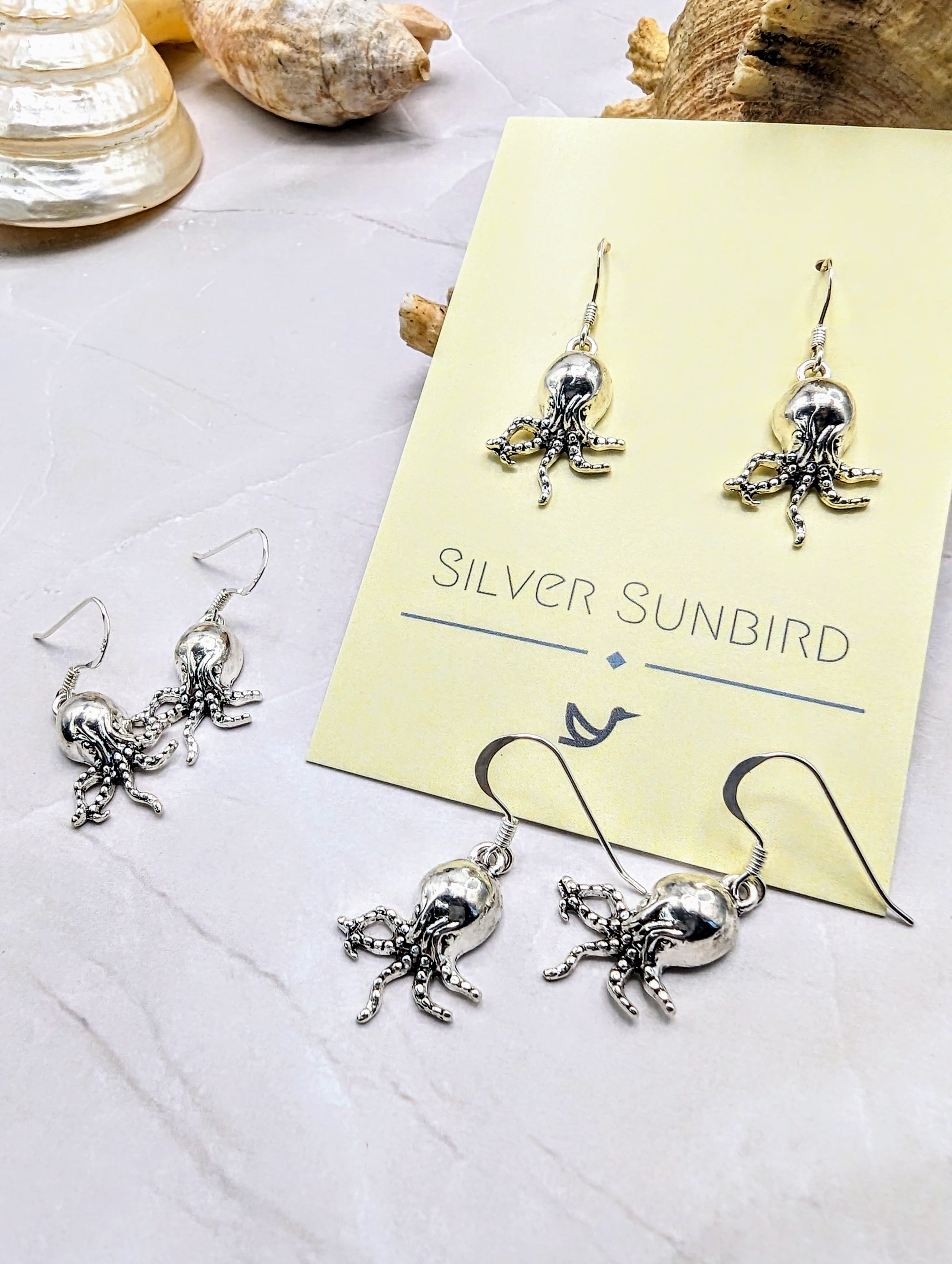 A display of Awesome Octopus Earrings is shown. Three different pairs of these intricate, Sterling Silver earrings are placed on a light-colored surface. Two pairs rest on a card labelled "Silver Sunbird," and seashell decorations in the background enhance the Ocean Jewellery theme.