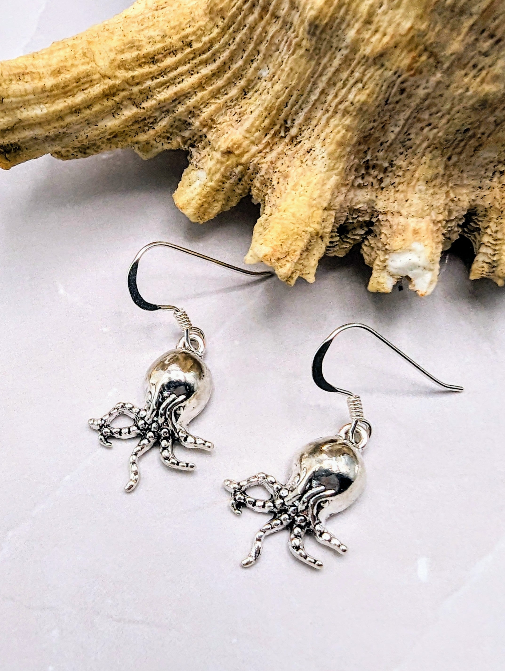A pair of Awesome Octopus Earrings by Silver Sunbird, made from sterling silver and featuring intricate detailing on the tentacles, resting on a textured beige seashell background. These hook earrings create a nautical theme perfect for any ocean jewellery collection.