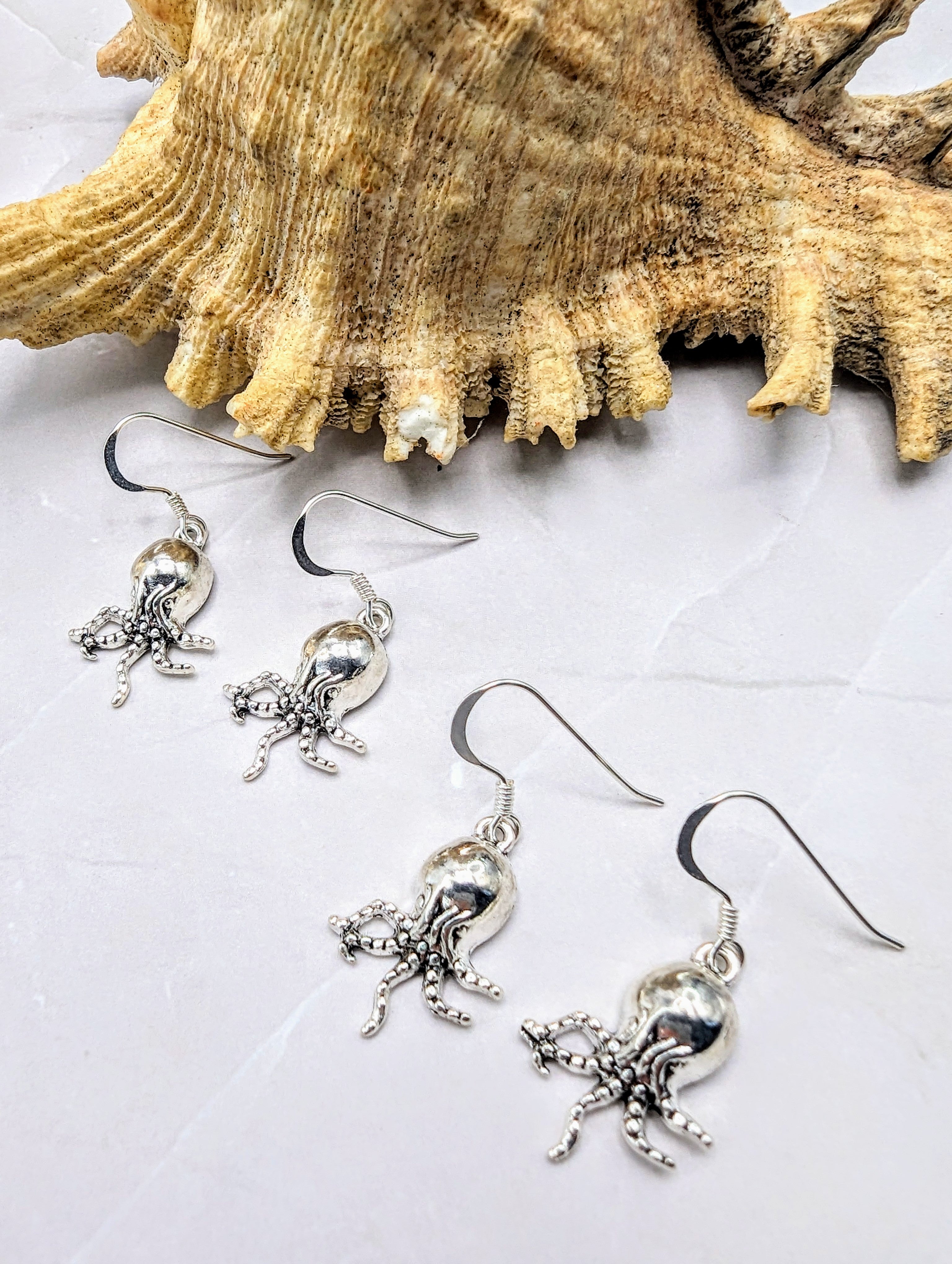 The Awesome Octopus Earrings by Silver Sunbird are sterling silver treasures displayed on a light-coloured marble surface with a large, textured seashell in the background. These exquisite pieces of ocean jewellery feature detailed tentacles and bodies that reflect light, creating a shiny appearance.