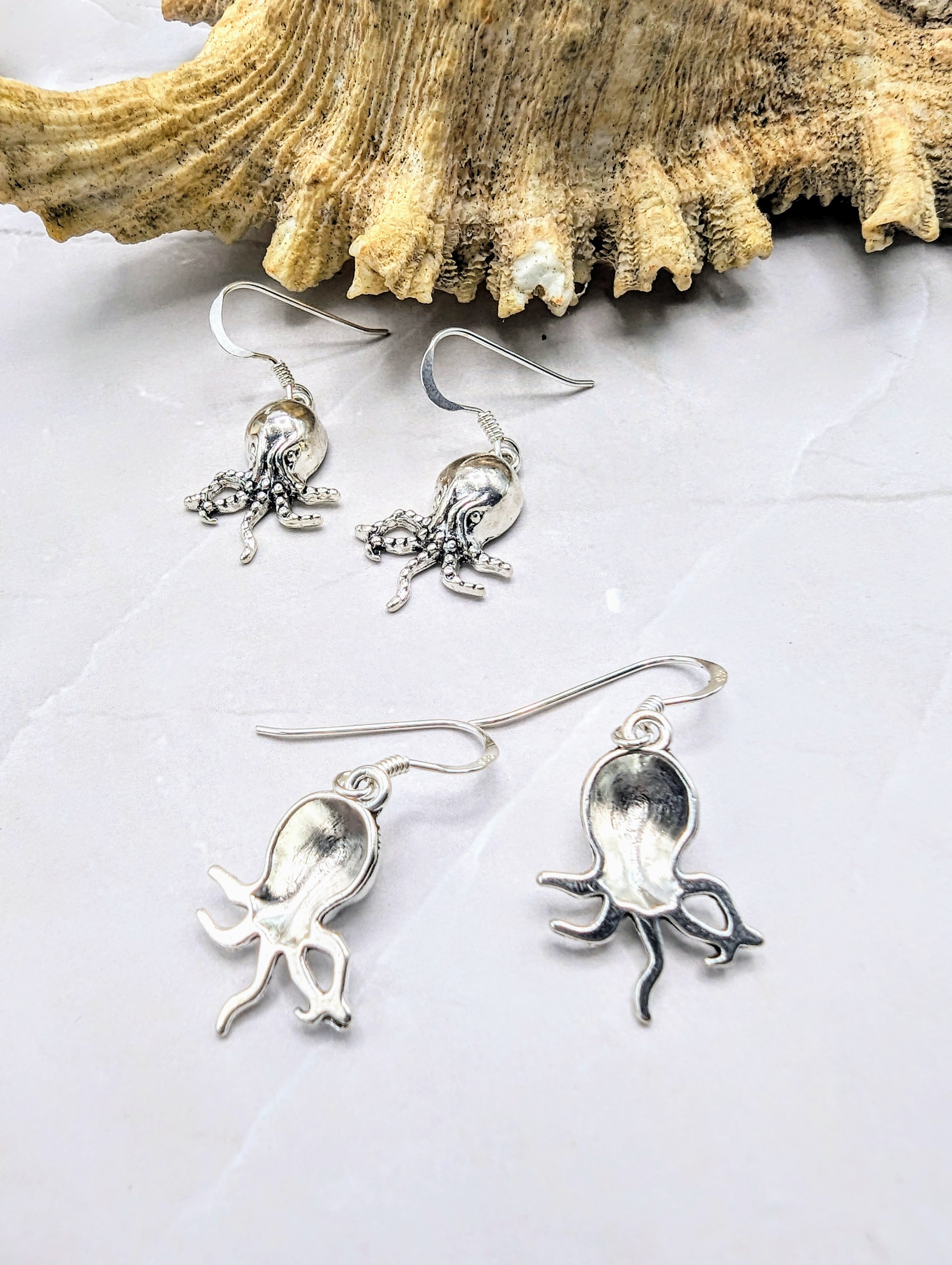 The Awesome Octopus Earrings by Silver Sunbird are displayed against a light gray stone surface. A decorative piece of coral reef shell is placed behind the earrings, enhancing the oceanic theme of this unique ocean jewellery.