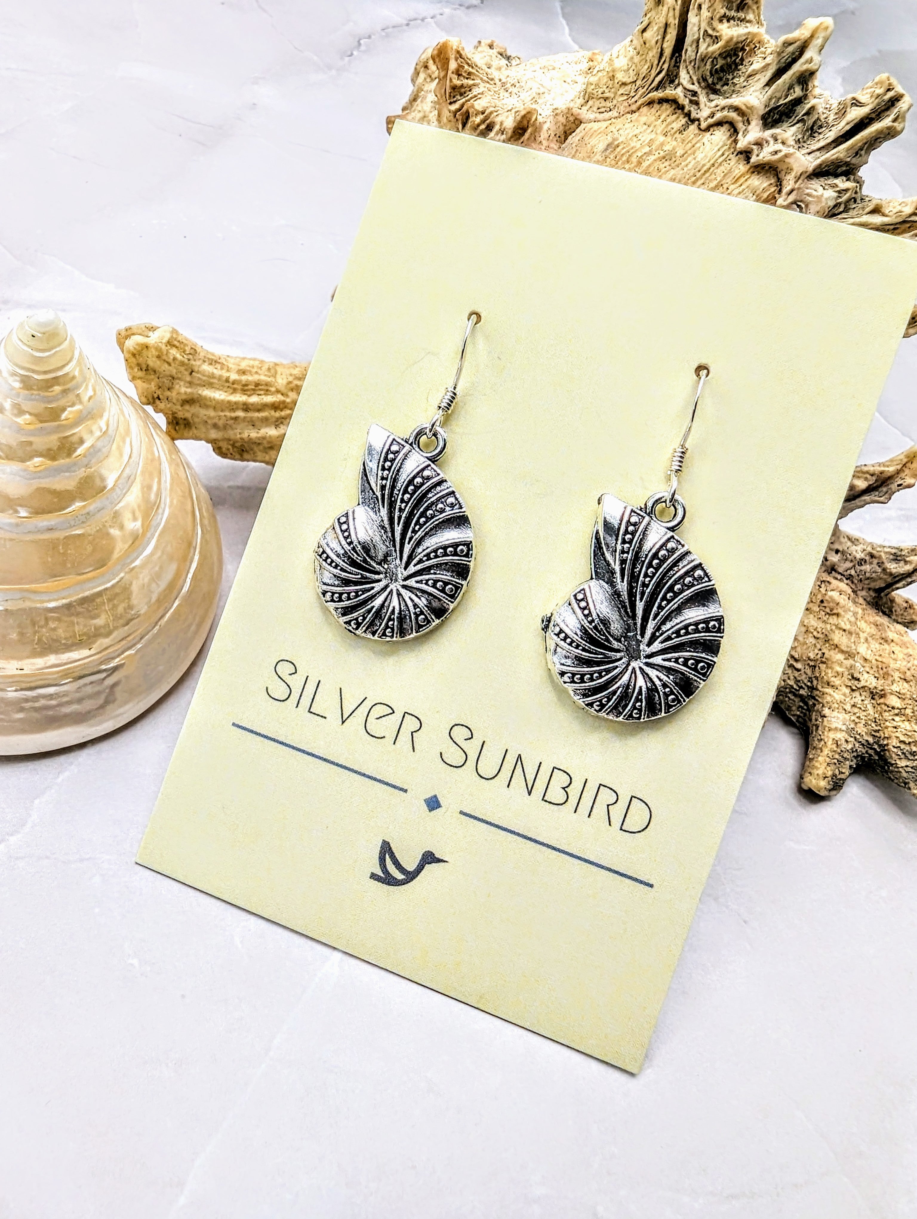 Shoreline Spiral Shell Earrings by Silver Sunbird, crafted from 925 sterling silver with hypoallergenic hooks, are shown on a branded card with seashells and a light stone background.