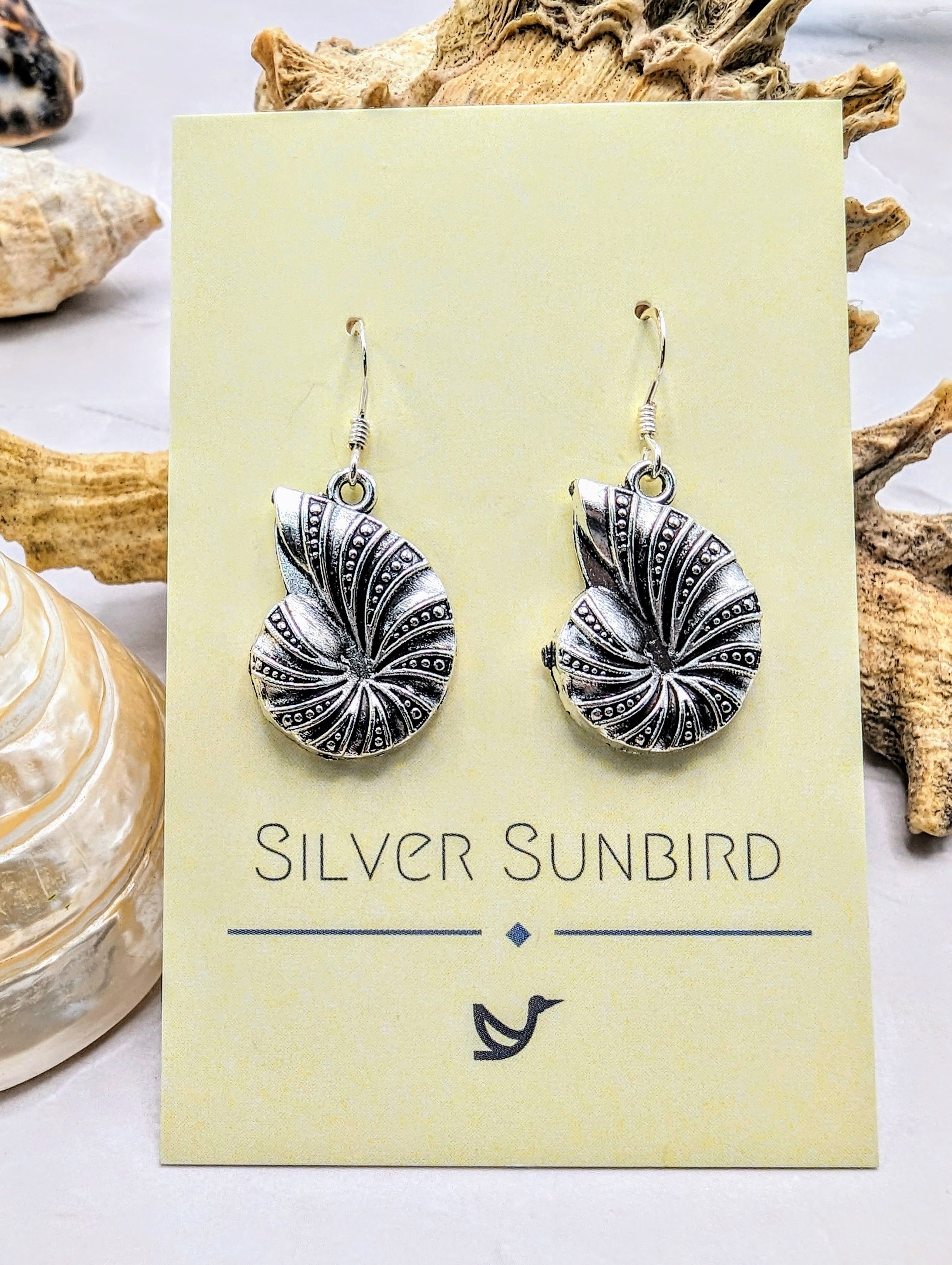 A pair of Shoreline Spiral Shell Earrings by Silver Sunbird, crafted from ornate 925 sterling silver with intricate shell-shaped detailing, displayed on a branded card and featuring hypoallergenic hooks for comfortable wear.