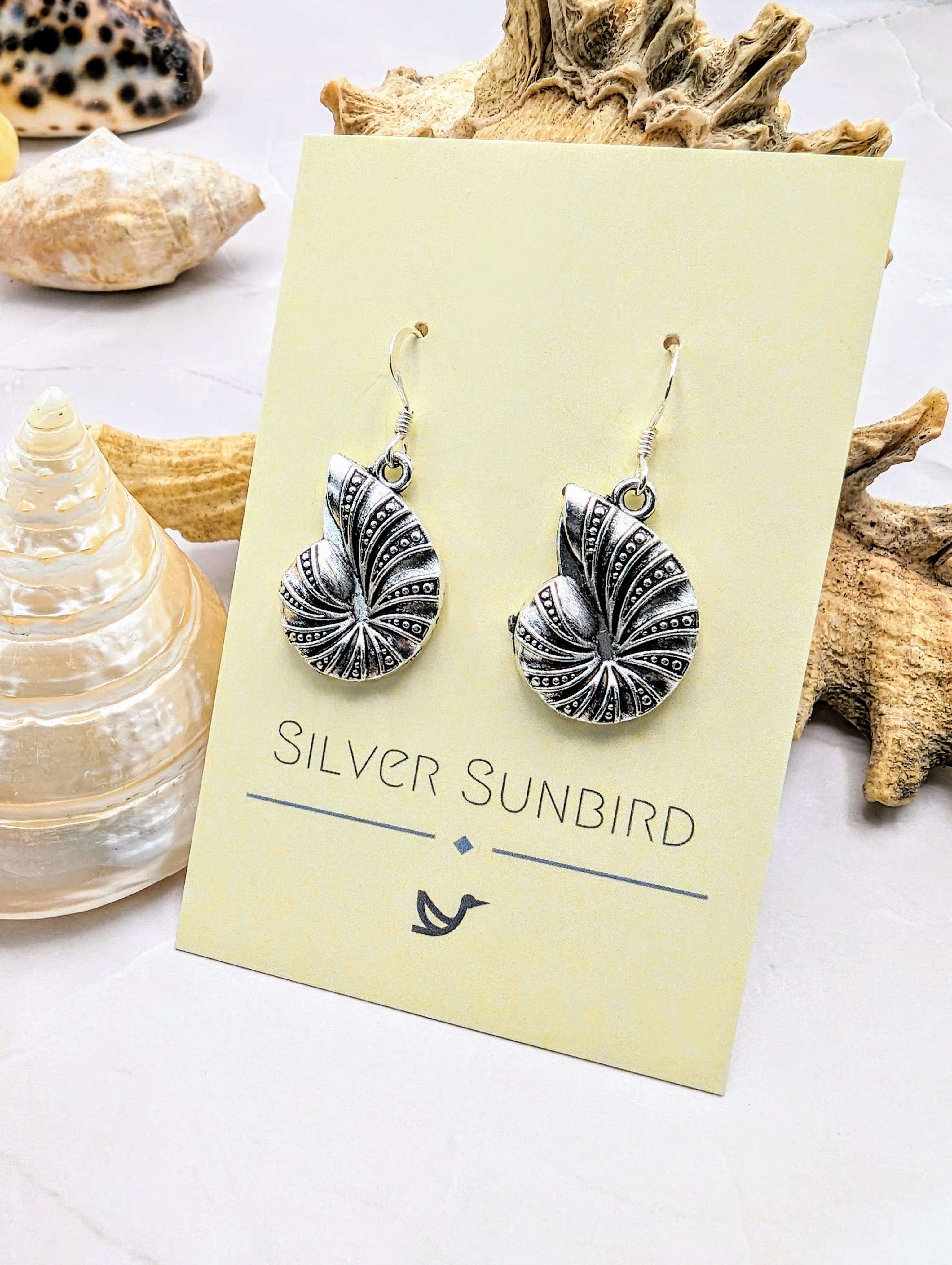The Shoreline Spiral Shell Earrings by Silver Sunbird feature intricate silver spirals and hypoallergenic sterling silver hooks, displayed on a branded card amid seashells and coral on a light surface.