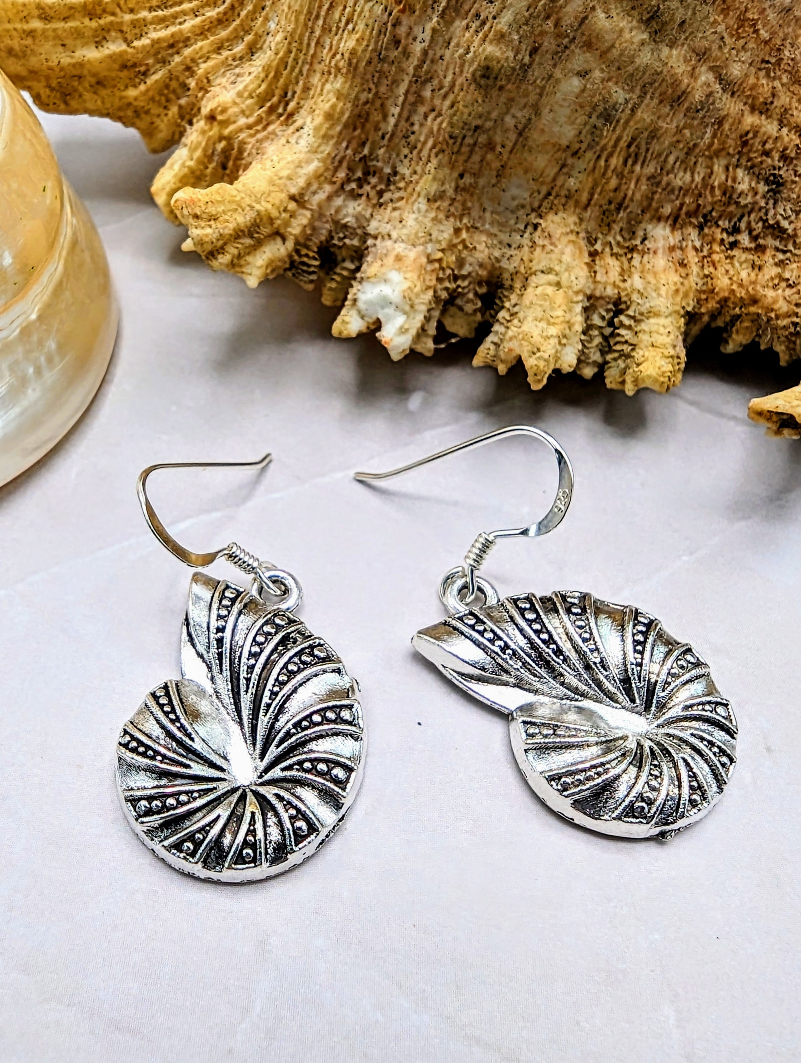 The Shoreline Spiral Shell Earrings by Silver Sunbird feature 925 sterling silver with intricate patterns and hypoallergenic hooks, displayed on a light surface near a large natural seashell.