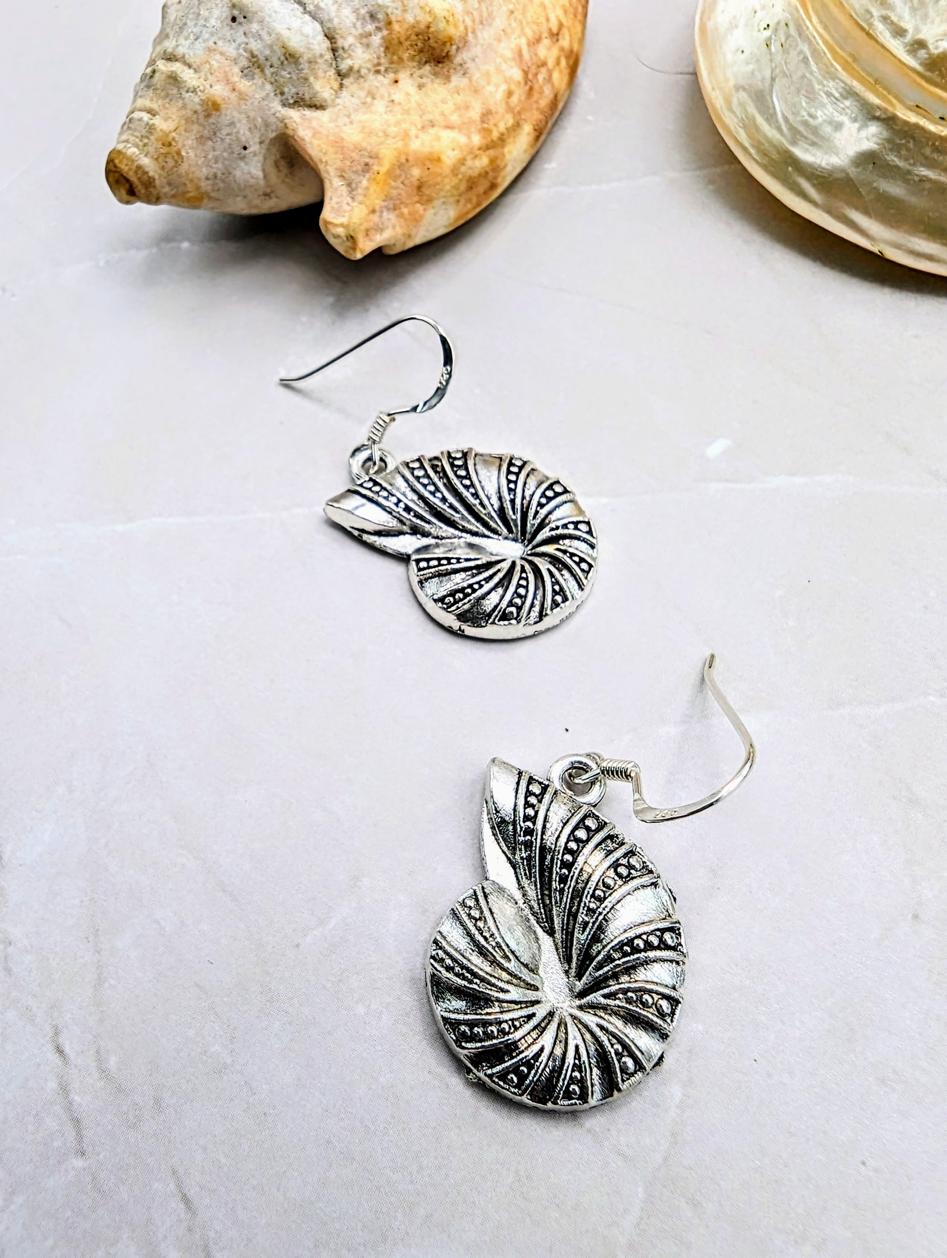 The Shoreline Spiral Shell Earrings by Silver Sunbird, featuring intricate detailing and hypoallergenic sterling silver hooks, are displayed on a light surface beside two seashells.