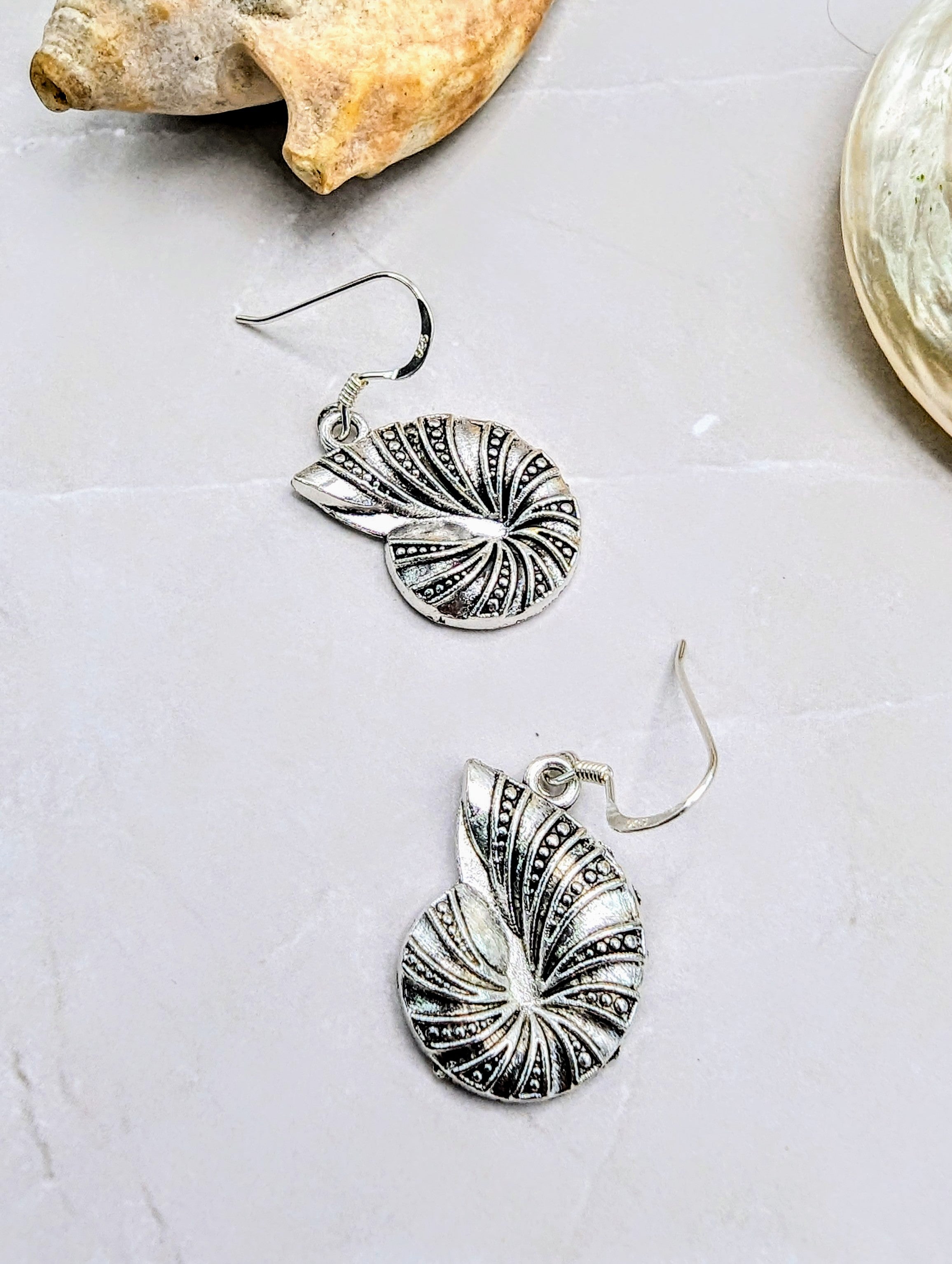 The Shoreline Spiral Shell Earrings by Silver Sunbird feature intricate swirling patterns and hypoallergenic sterling silver hooks. Shown on a light stone surface beside two natural seashells, these earrings evoke seaside elegance.