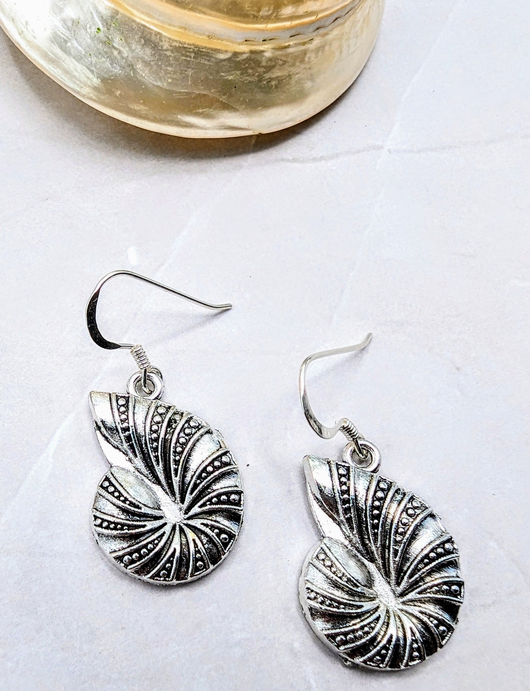 Silver Sunbird’s Shoreline Spiral Shell Earrings, made from 925 sterling silver with intricate dot patterns, are displayed on a white surface beside a polished translucent shell.
